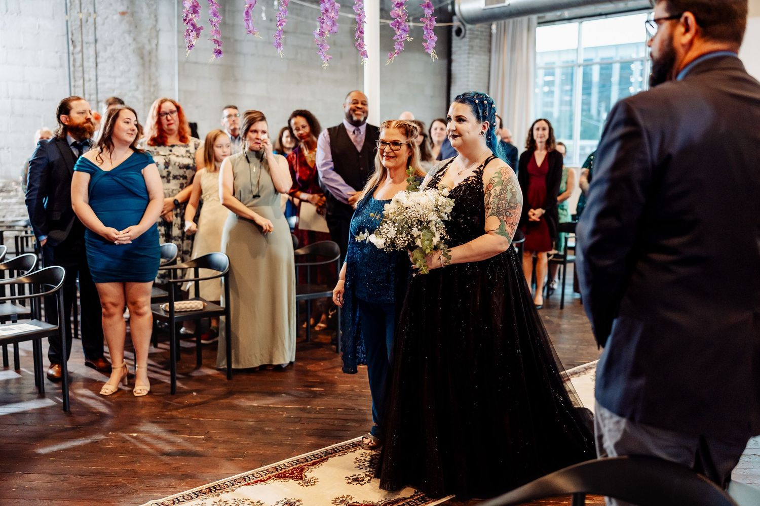 Tattooed bride in black gown walks down aisle escorted by bridesmaid in navy dress.
