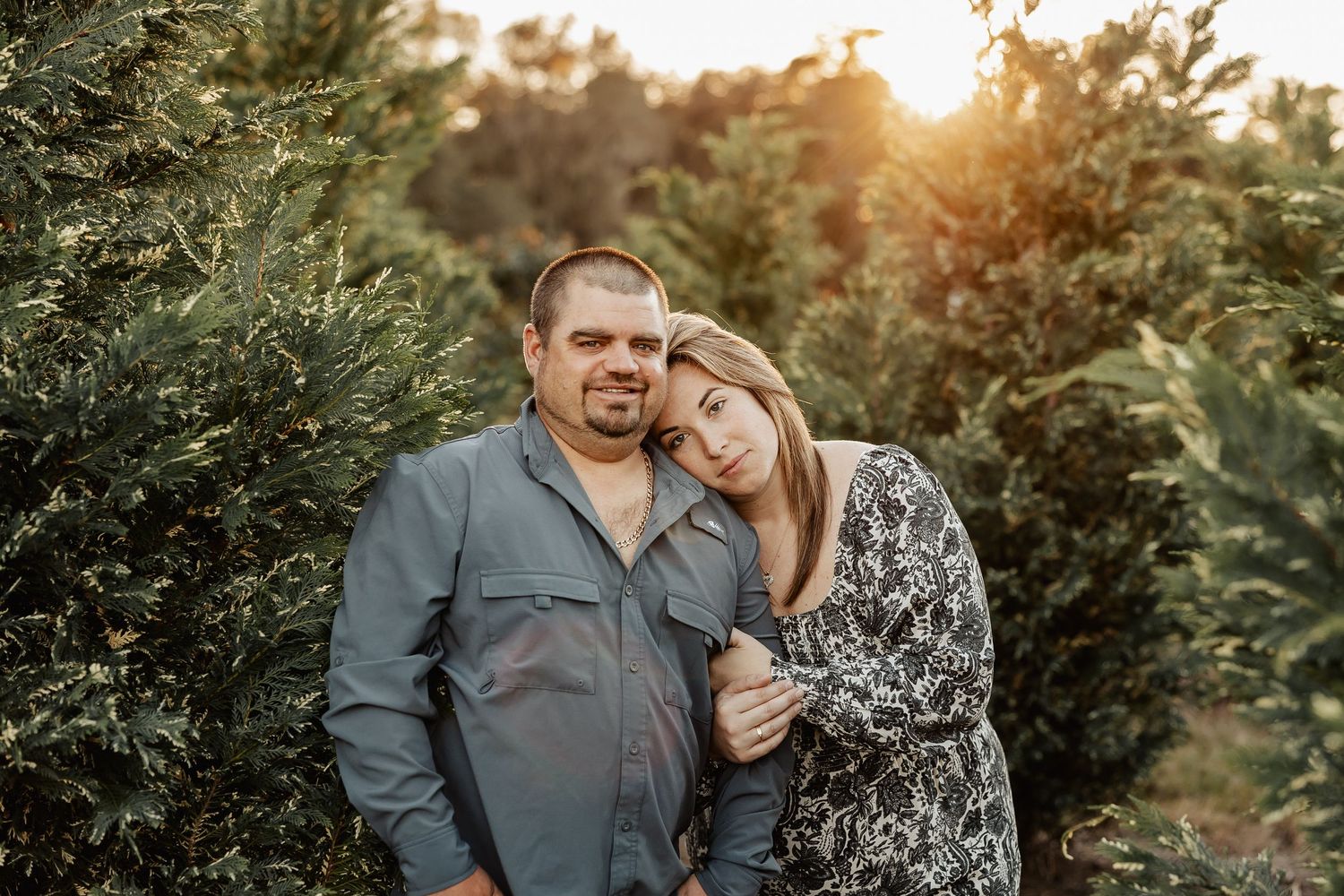 Couple embracing among lush green trees at golden hour, woman leaning on mans shoulder in floral dress.