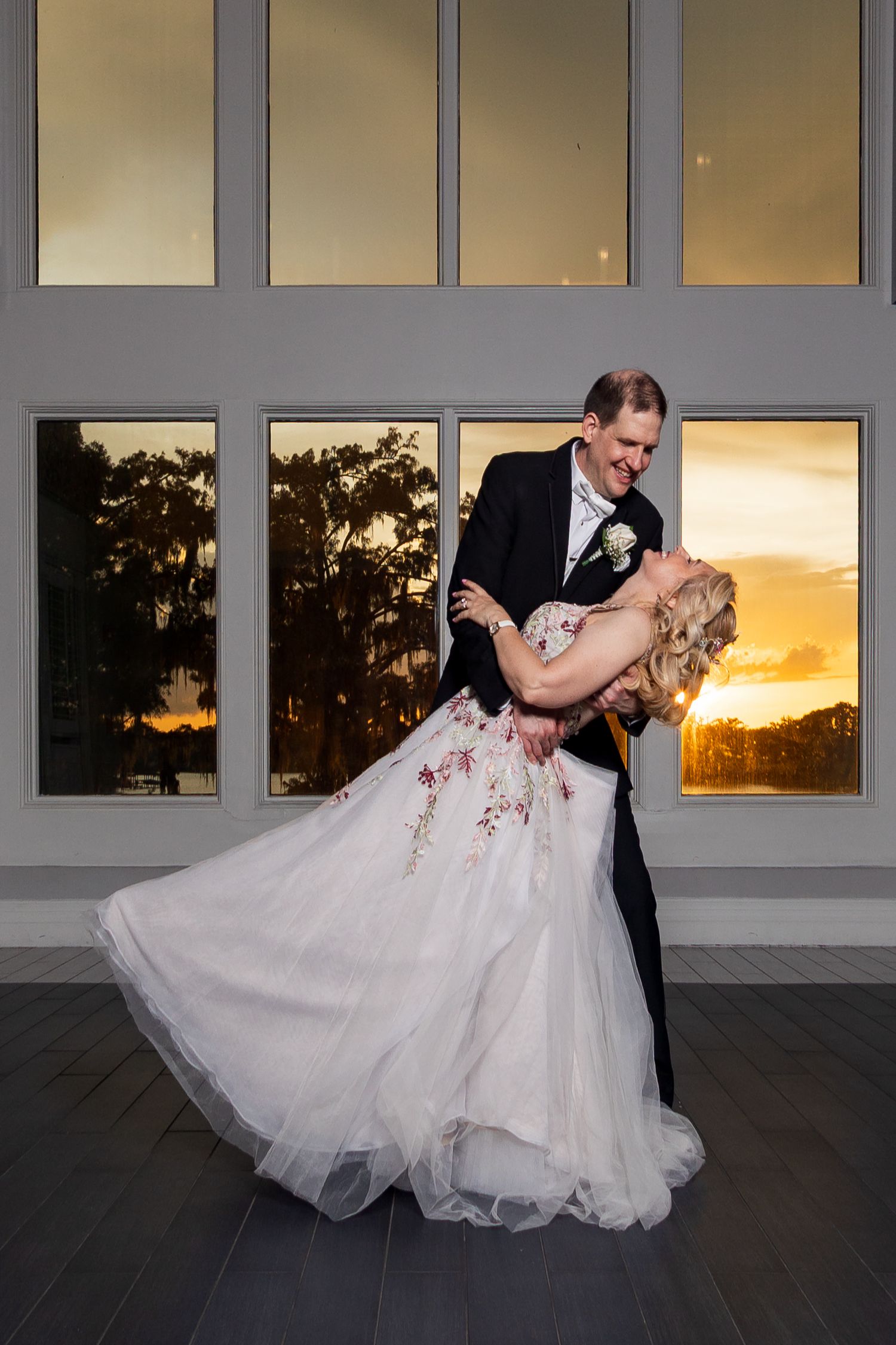Groom dipping bride in floral gown before large windows reflecting stunning golden sunset sky.