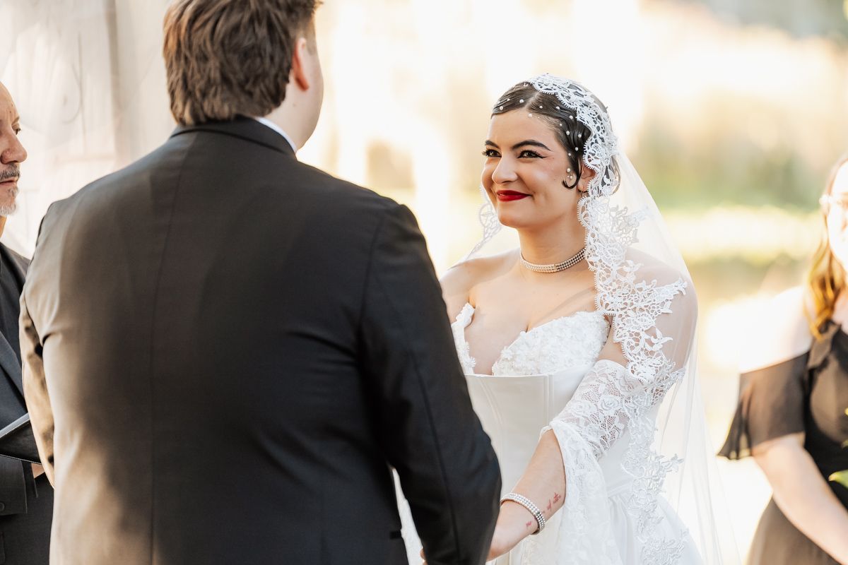 Smiling bride in lace mantilla veil with red lips holds grooms hands during outdoor ceremony.