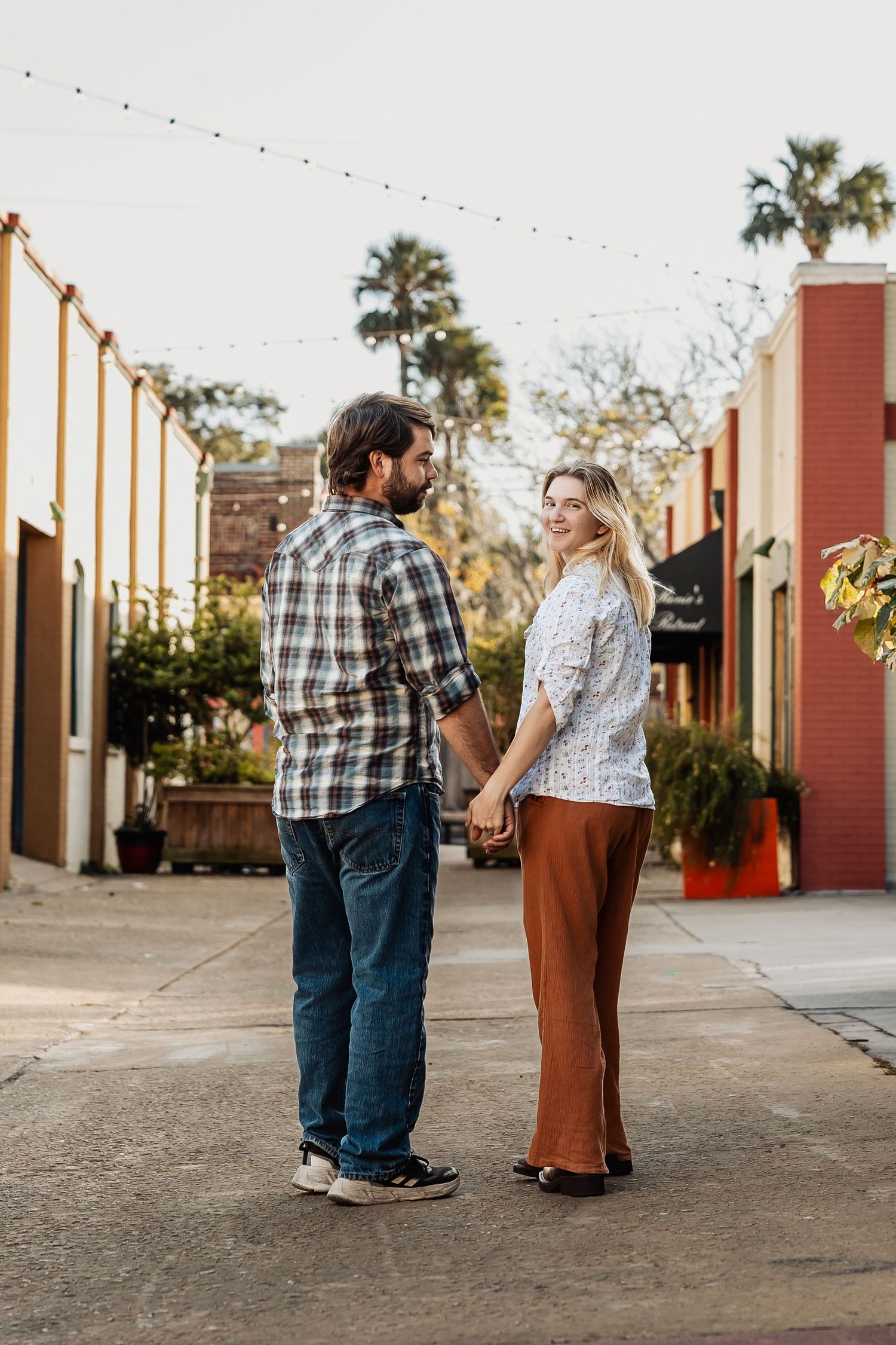 Couple holding hands on a charming outdoor walkway lined with string lights and palm trees.