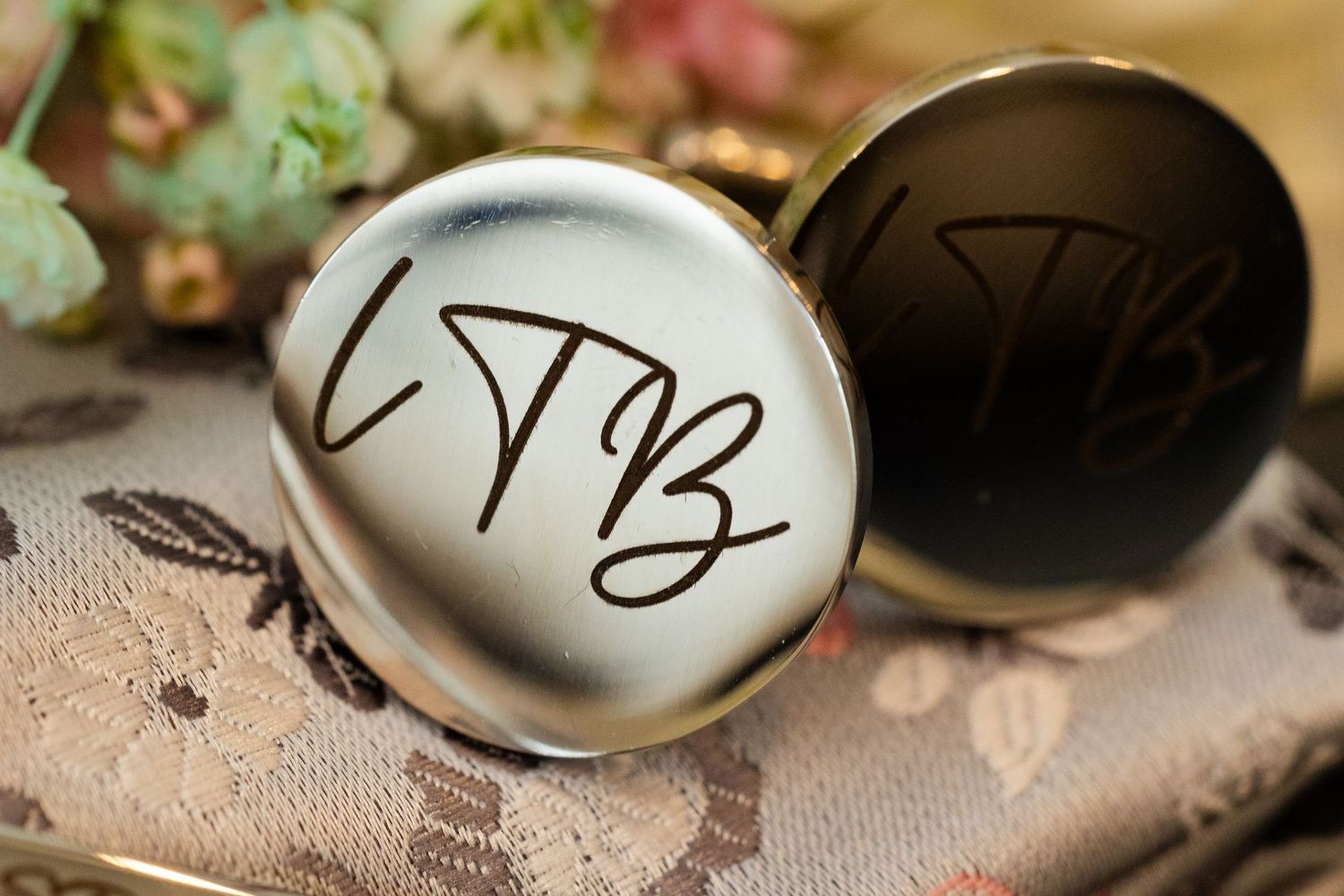 Close-up of two custom cufflinks engraved with LTB initials in cursive, one silver and one black.