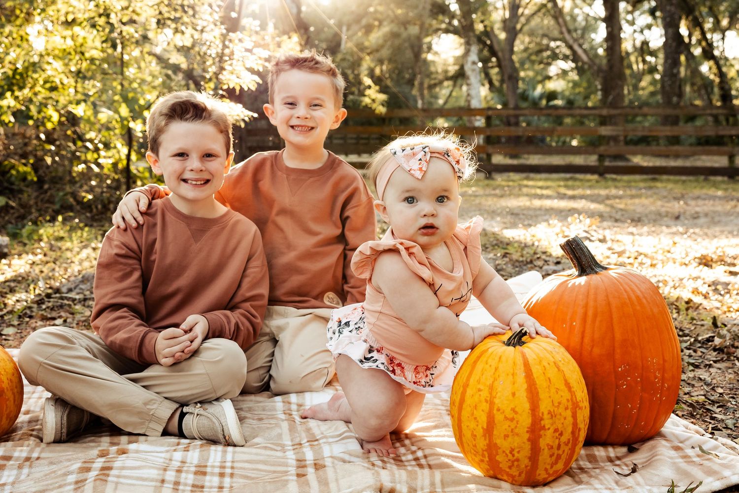 Three siblings sitting on a plaid blanket outdoors with orange pumpkins in a sunny fall setting.