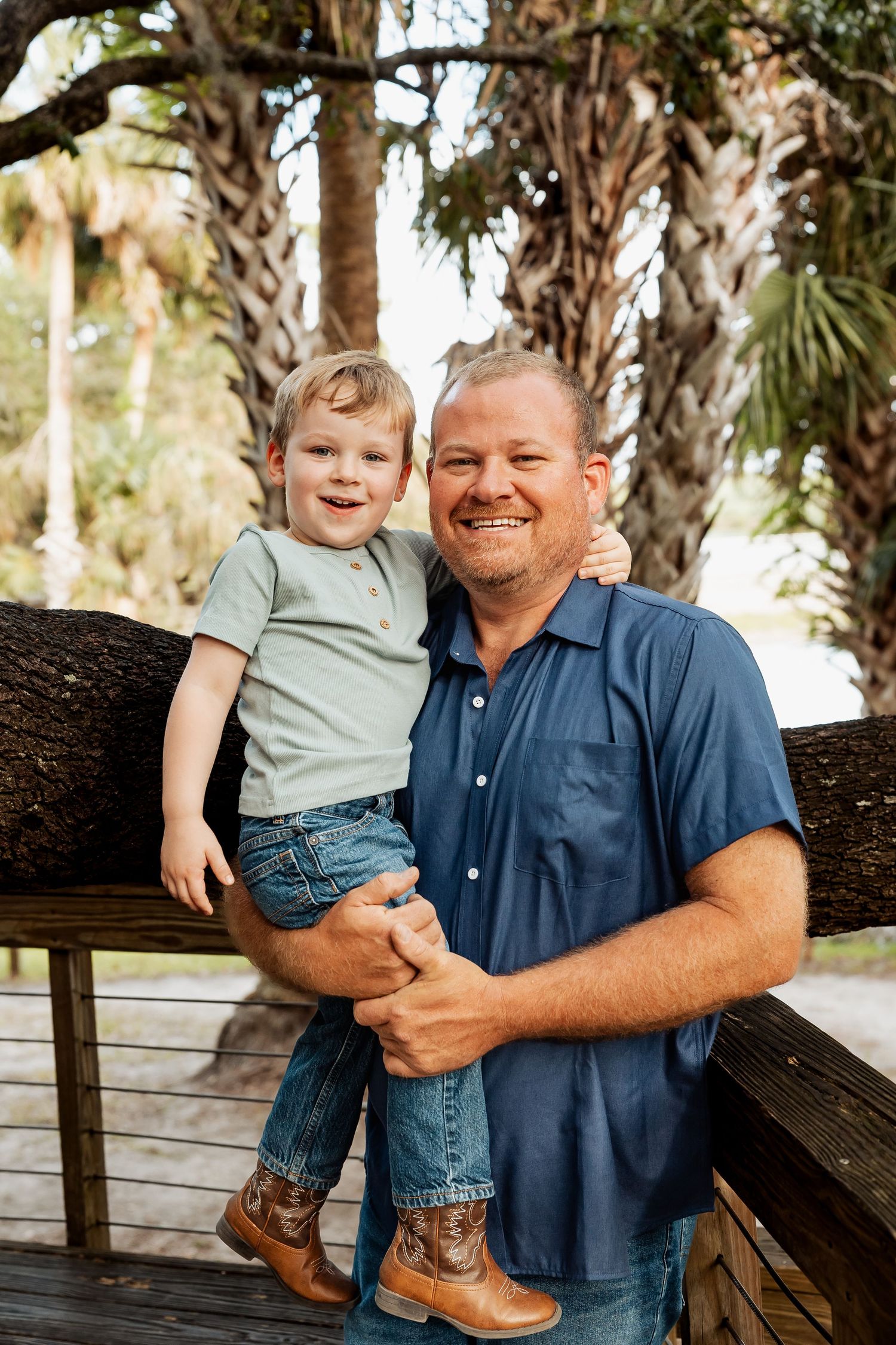 Smiling father holding his young toddler son outdoors near palm trees, both wearing casual blue outfits.