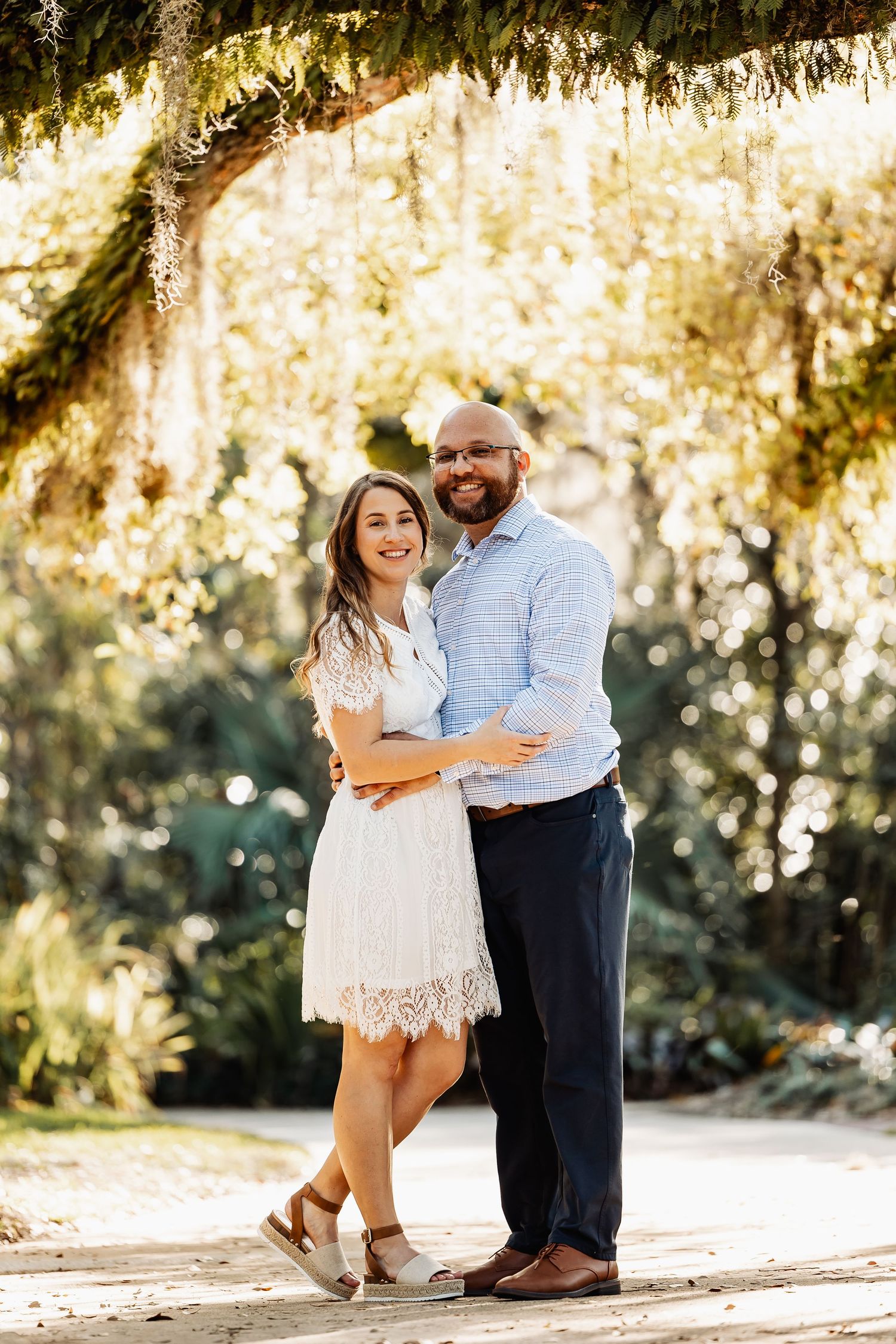 Couple embracing and smiling under canopy of Spanish moss-covered trees in golden afternoon light.