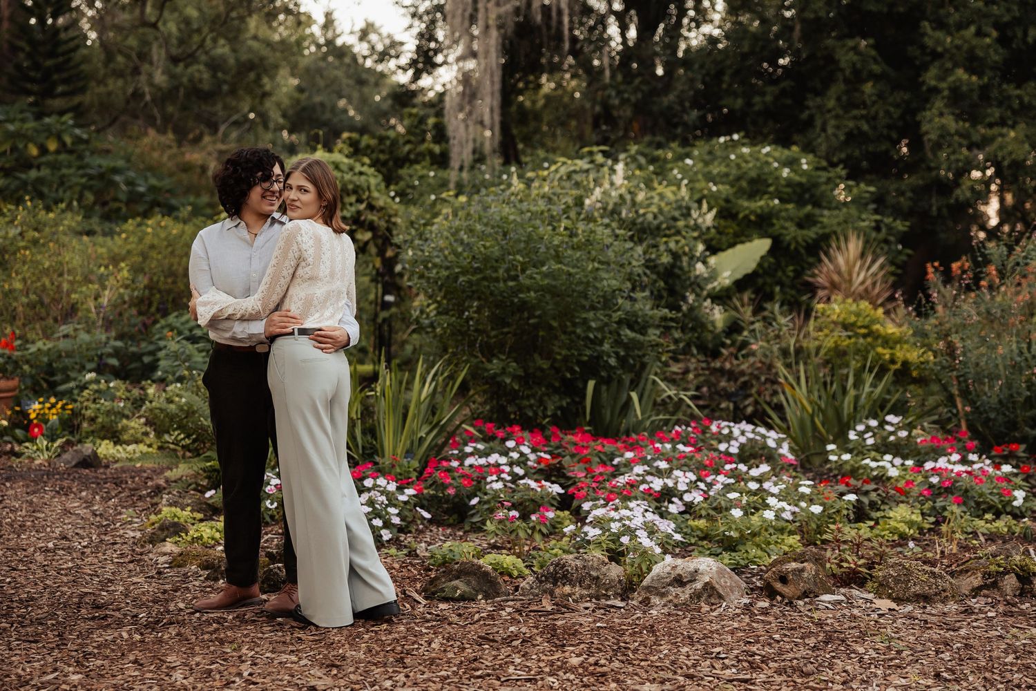 Two women embracing in a lush garden surrounded by colorful flowers and greenery.