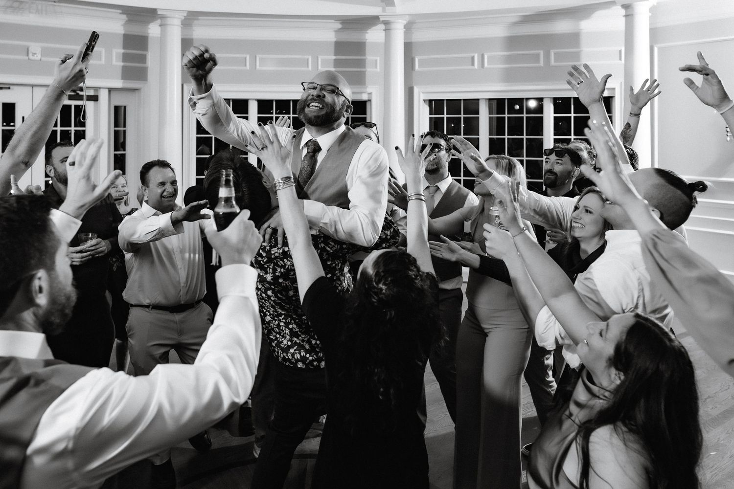 Groom holds bottle aloft surrounded by cheering guests in black and white wedding reception photo.