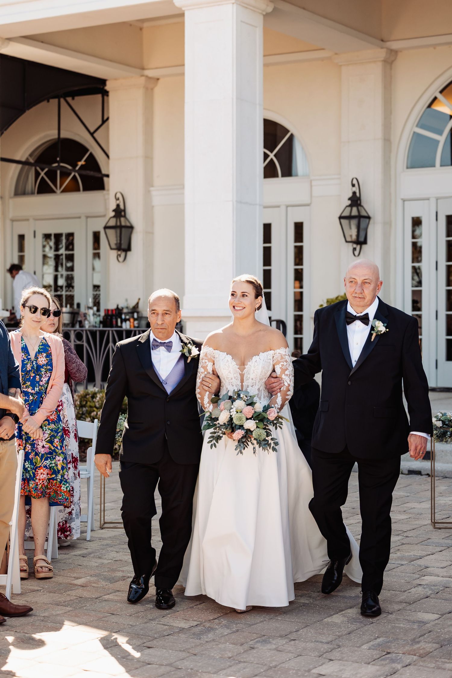 Smiling bride in lace gown walks down the aisle escorted by two men in black tuxedos holding bouquet.