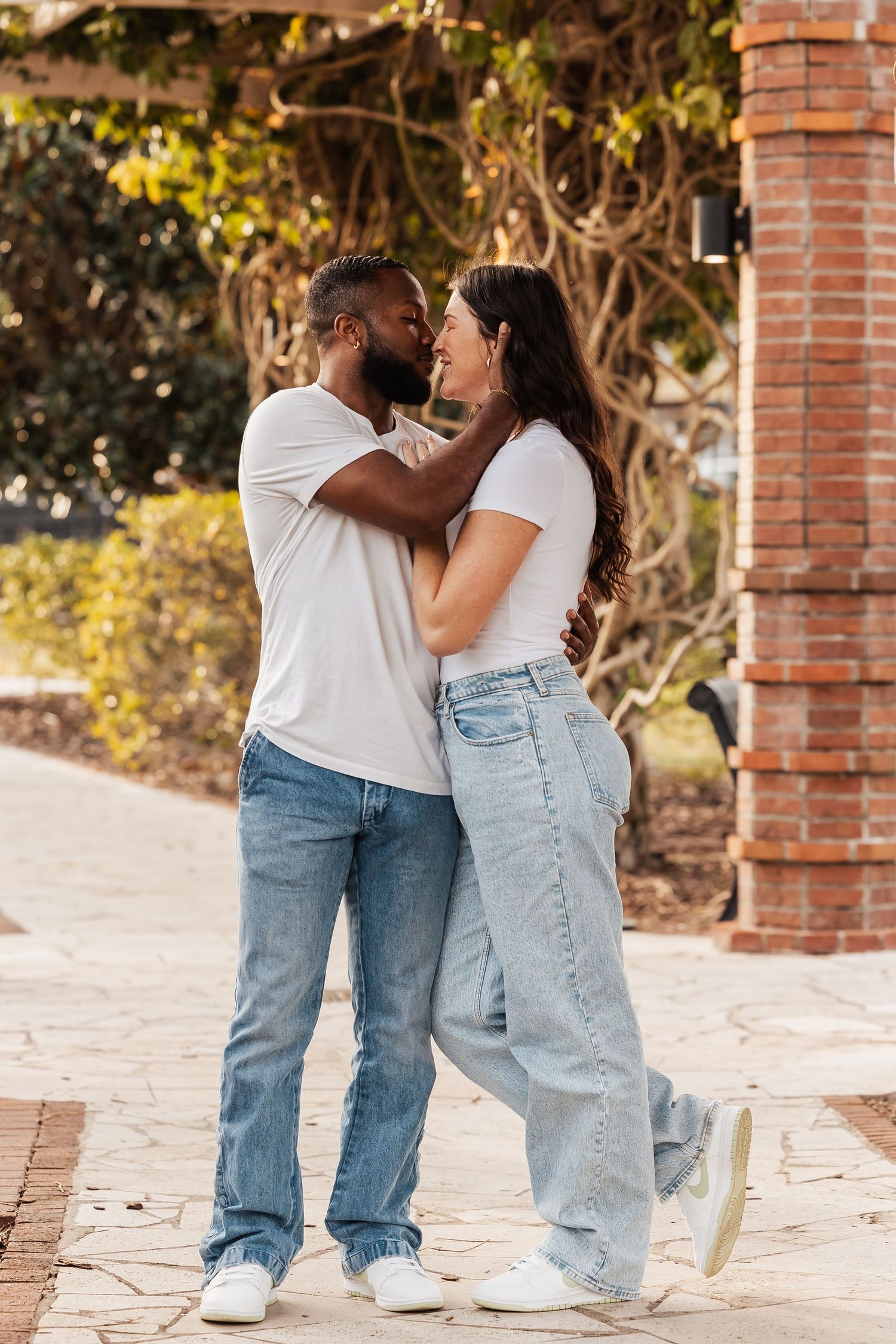 Loving couple in white shirts and jeans embracing face to face under green ivy archway outdoors.