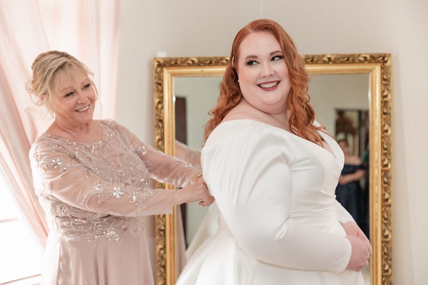 Mother helping red-haired bride into her elegant white long-sleeve wedding gown in front of a gold mirror.