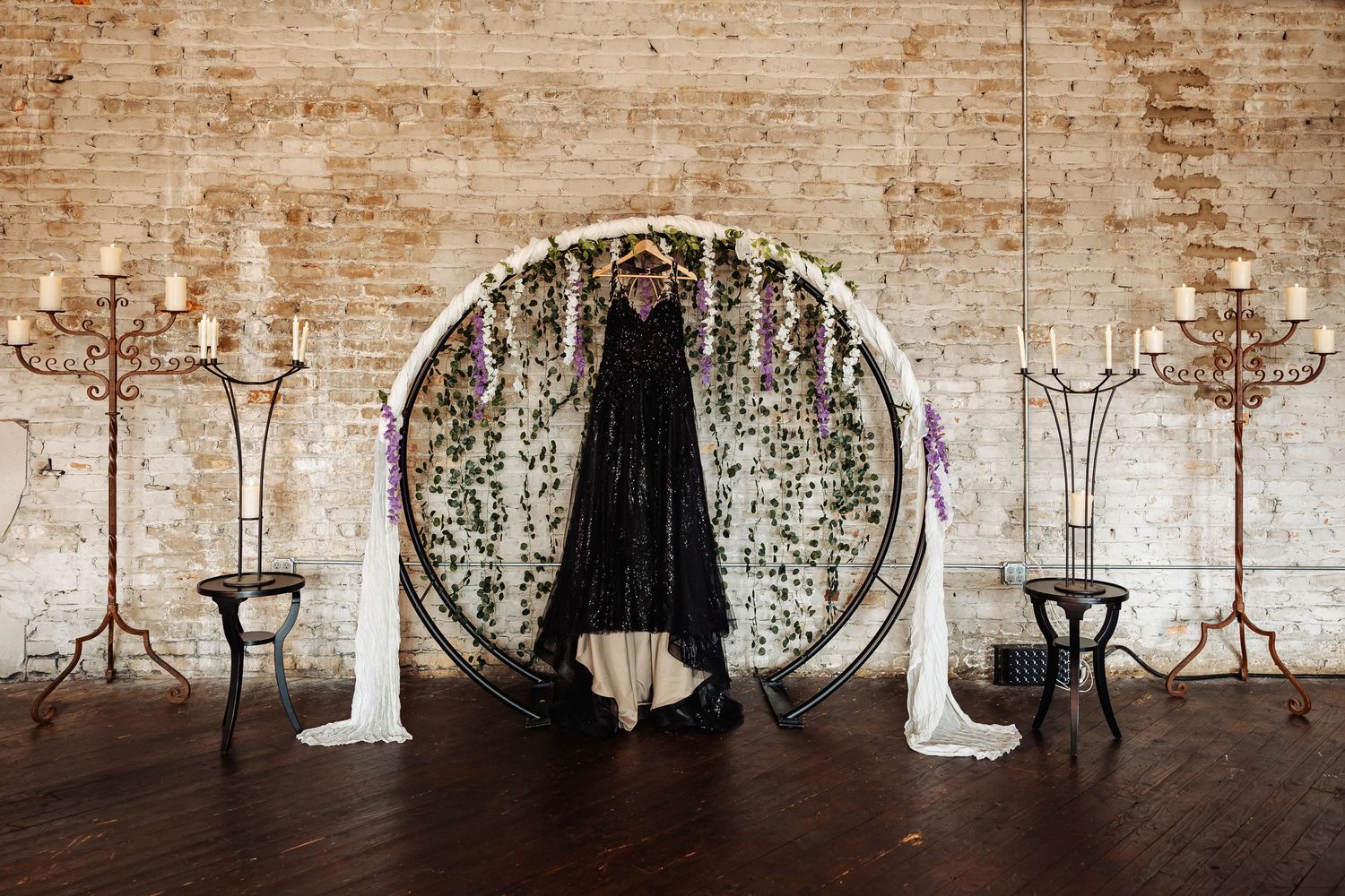 Black wedding gown displayed on floral circular arch with candelabras in rustic brick venue.