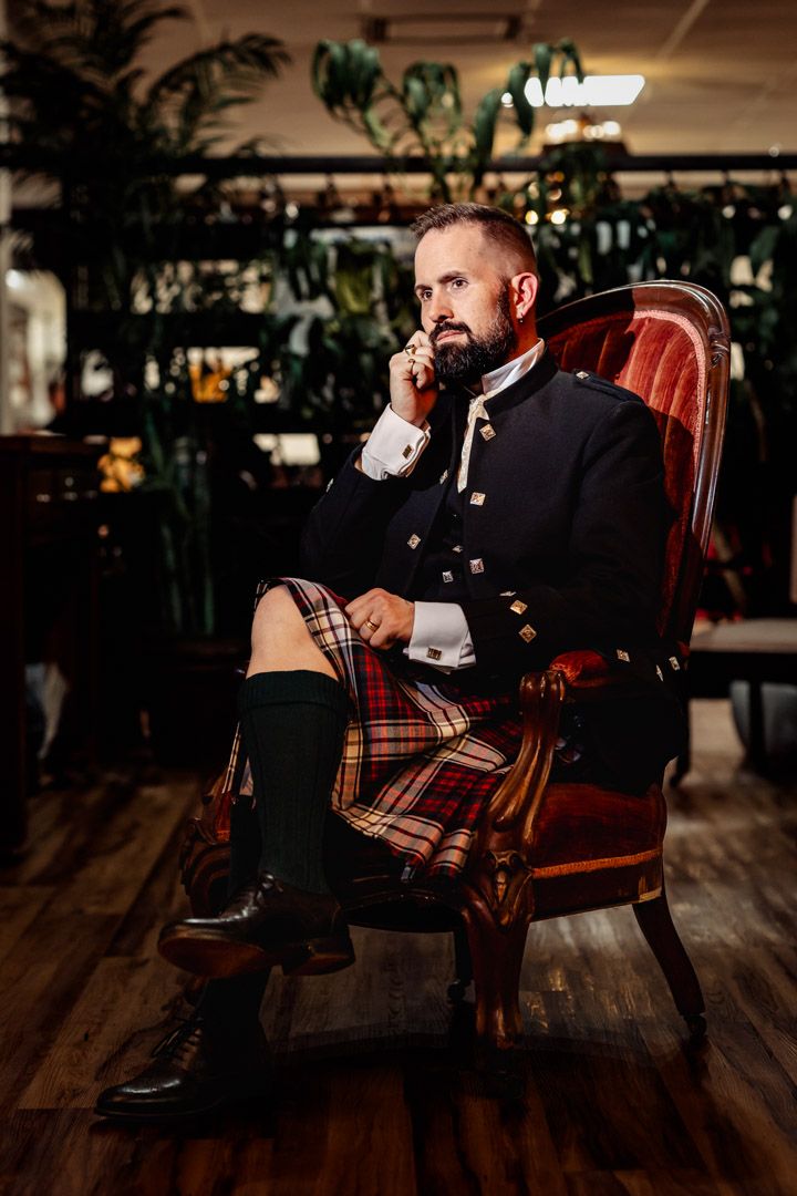 Bearded man in Scottish Highland dress sitting in velvet chair on phone.