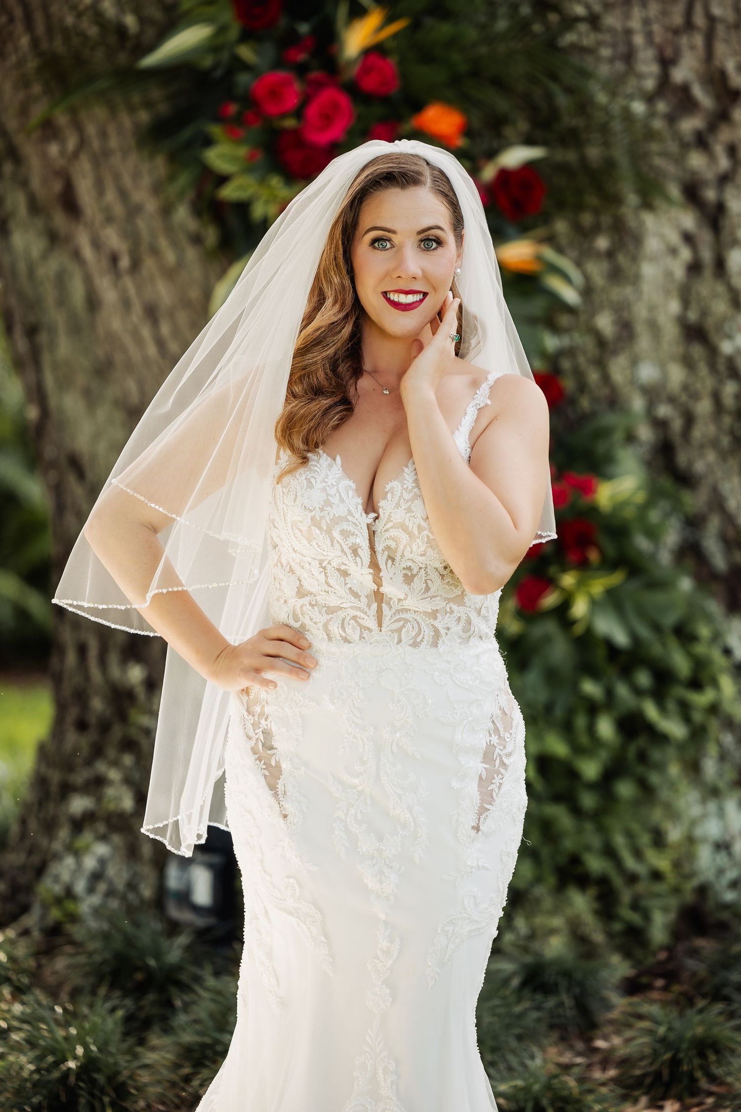 Elegant bride in lace mermaid gown with veil poses confidently before colorful floral backdrop outdoors.