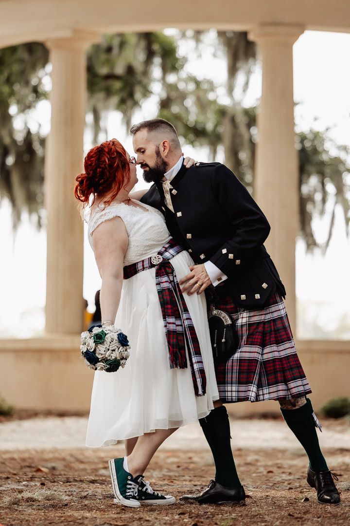 Bride with red hair holding bouquet and groom in Scottish kilt sharing a kiss near columned pavilion.