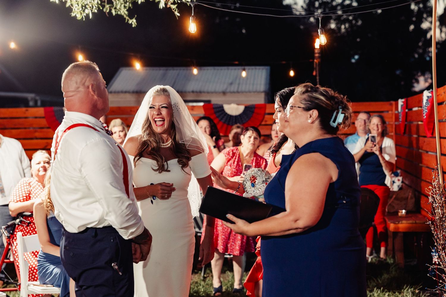 Bride laughing joyfully during outdoor wedding ceremony with string lights and officiant reading vows at night.