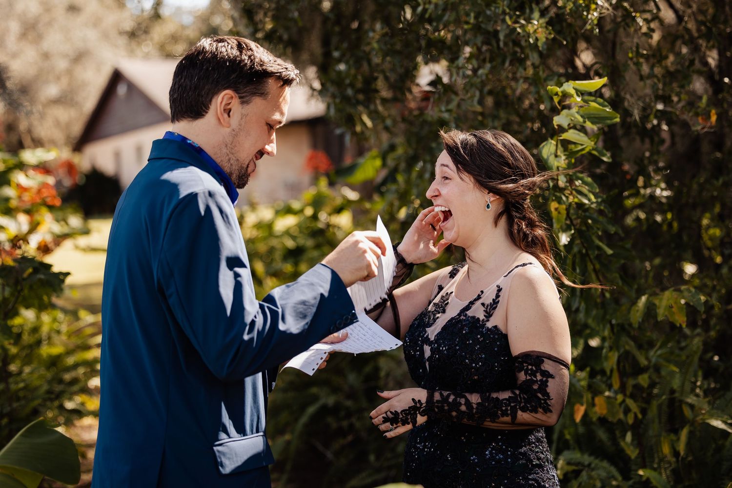 Groom reading vows to laughing bride in black lace dress during outdoor garden wedding ceremony.