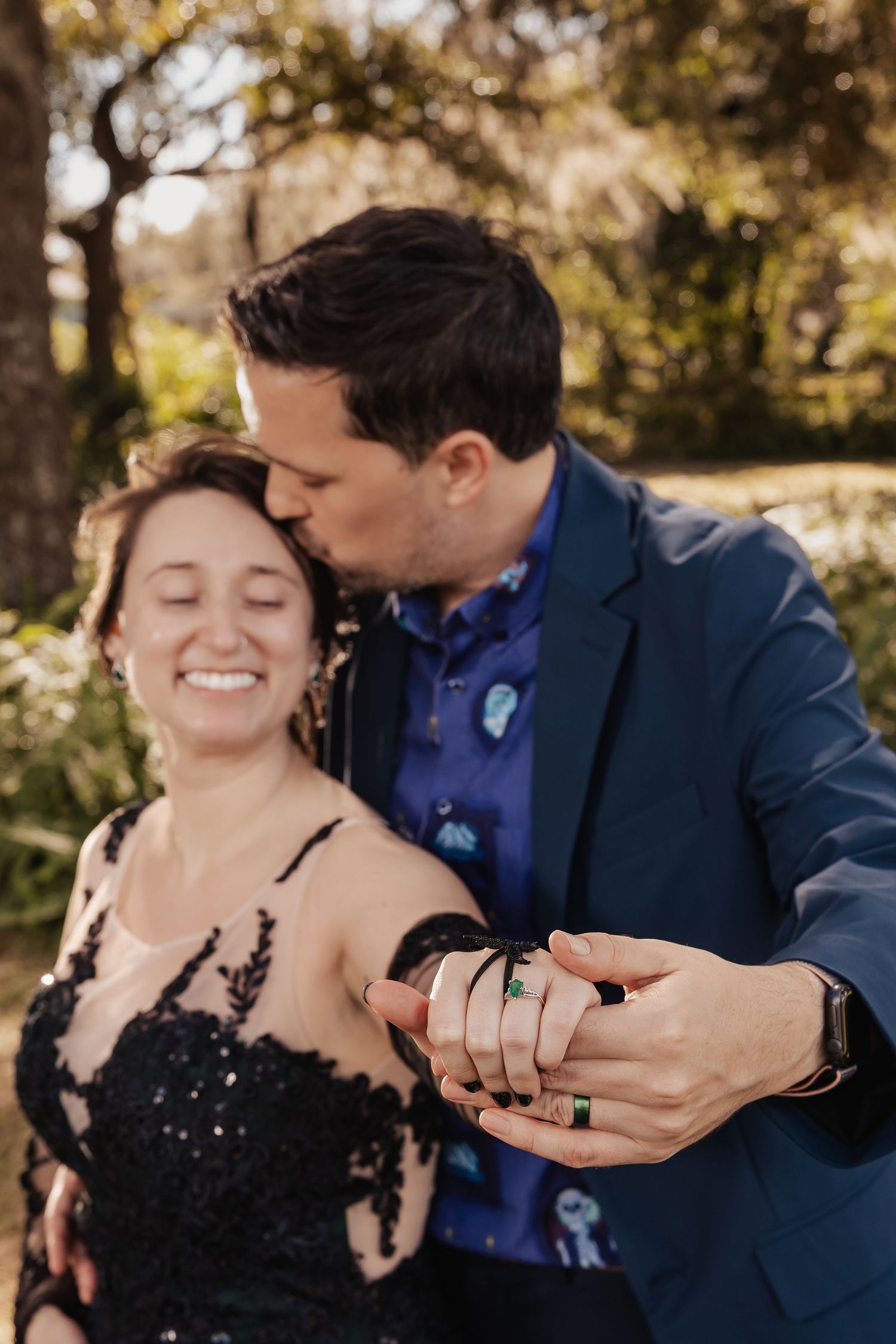 Smiling bride displays emerald wedding ring as groom kisses her head at romantic outdoor garden wedding.