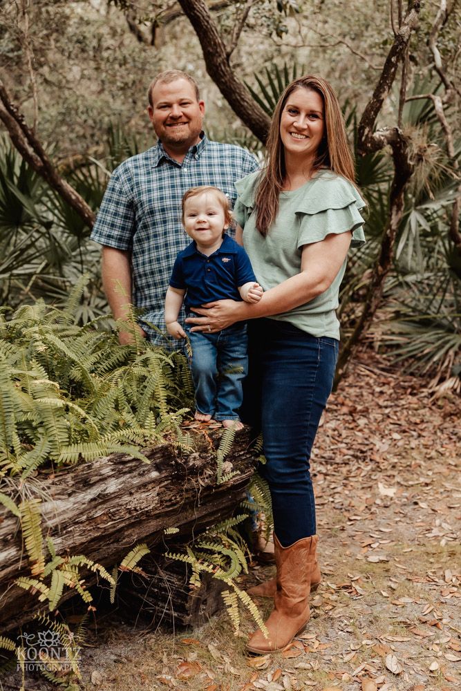 Family of three poses together on a fallen log surrounded by lush ferns and palm trees.