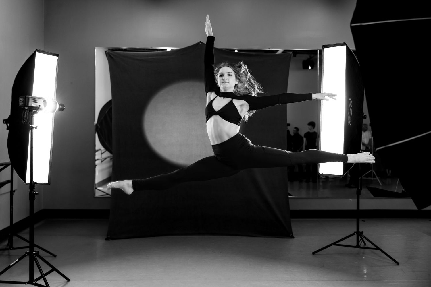 Dancer in black outfit performing dynamic leap in black and white studio photo.