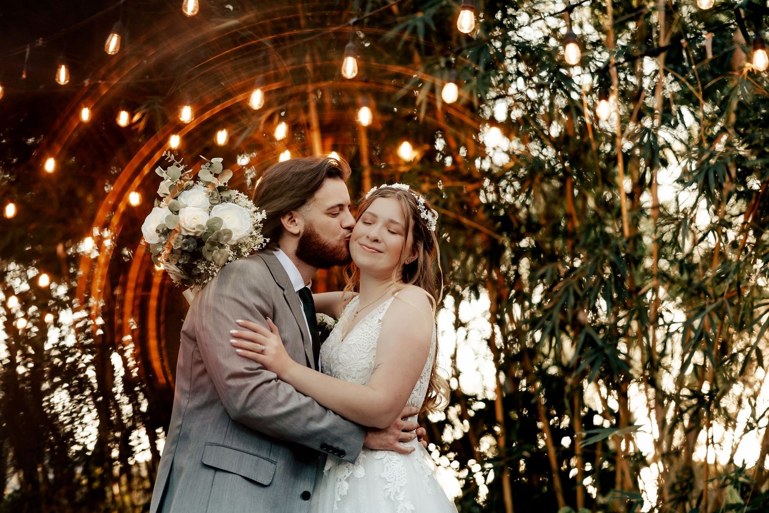 Wedding couple embraces in a magical garden setting with warm string lights and greenery as backdrop.