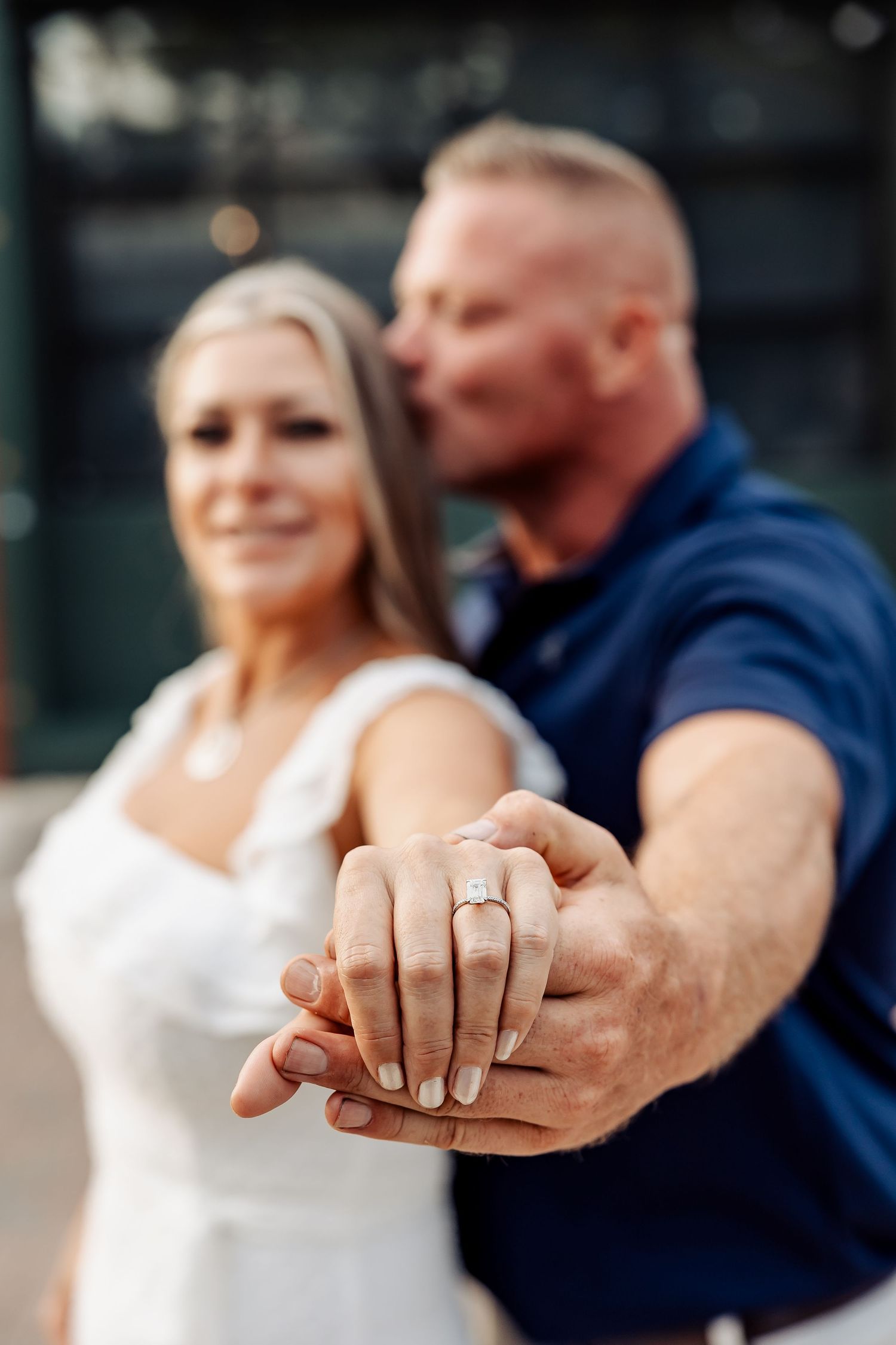 Engaged couple showing off diamond ring to camera, man kissing woman on cheek, soft focus background.