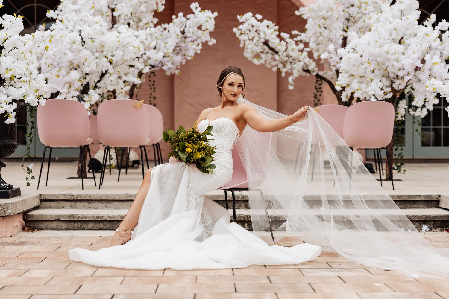 Elegant bridal portrait on terrace with flowing veil and white cherry blossom trees against pink backdrop.