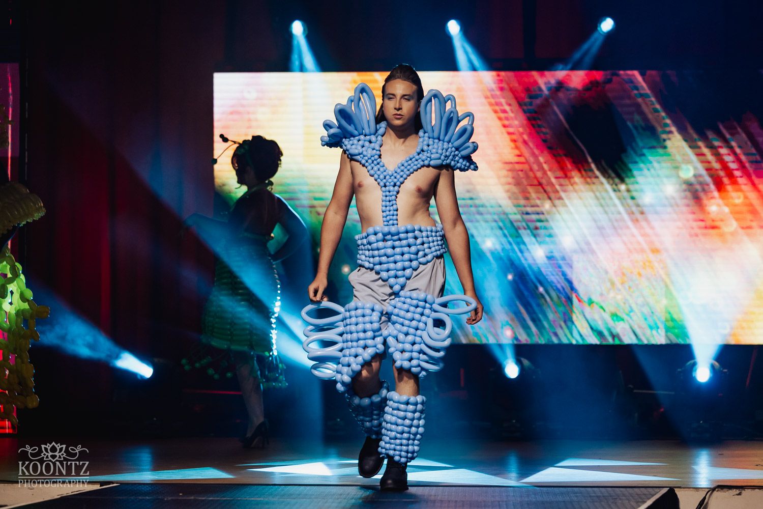 Dancer performs energetic moves on dark stage with colorful light effects and spotlights creating dramatic atmosphere.