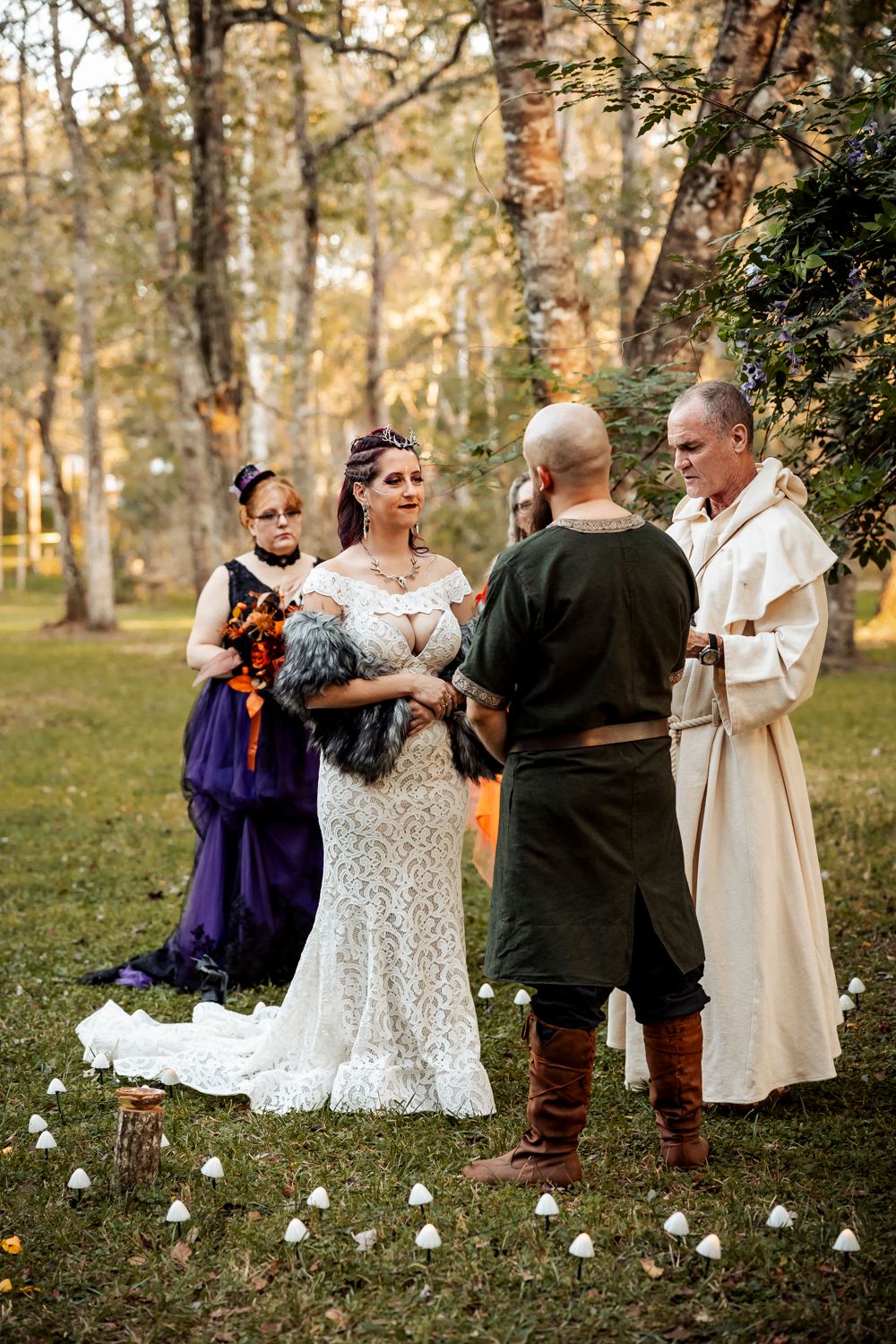 Medieval-themed outdoor wedding ceremony with couple and clergy in period costumes beneath autumn trees.