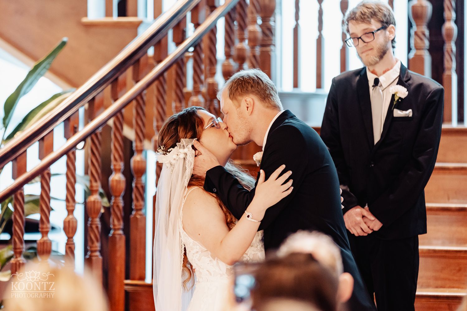 A romantic kiss during a wedding ceremony on elegant wooden stairs with onlookers in attendance.