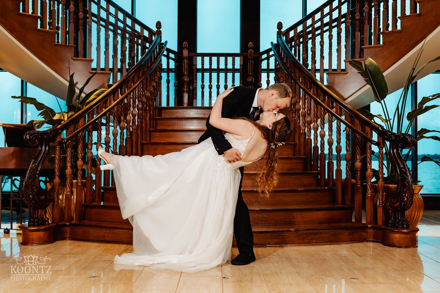 Dramatic wedding dip pose on grand wooden staircase with indoor plants.