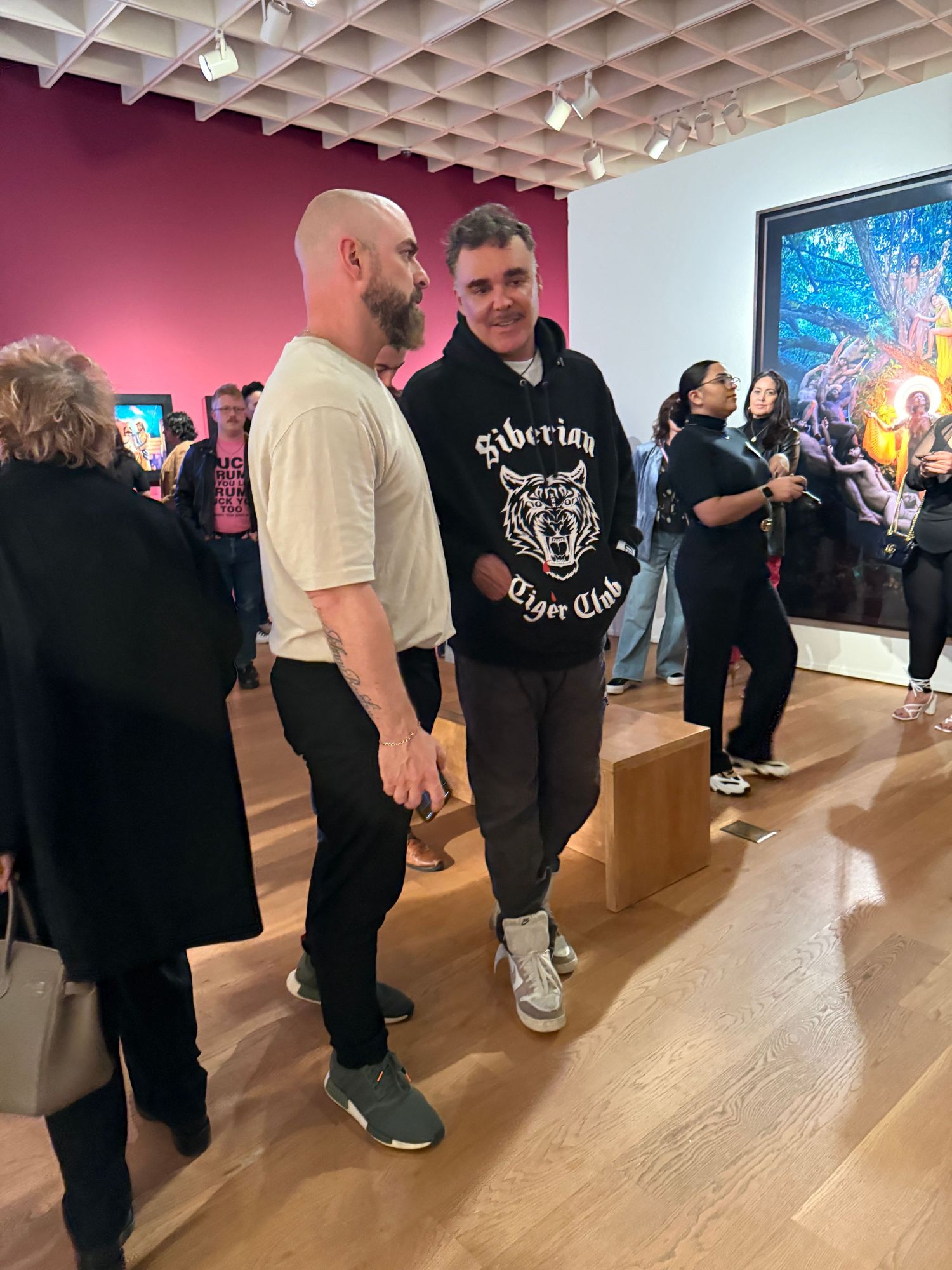 A group of people standing in an art gallery with wooden floors and white walls viewing colorful artwork on display.