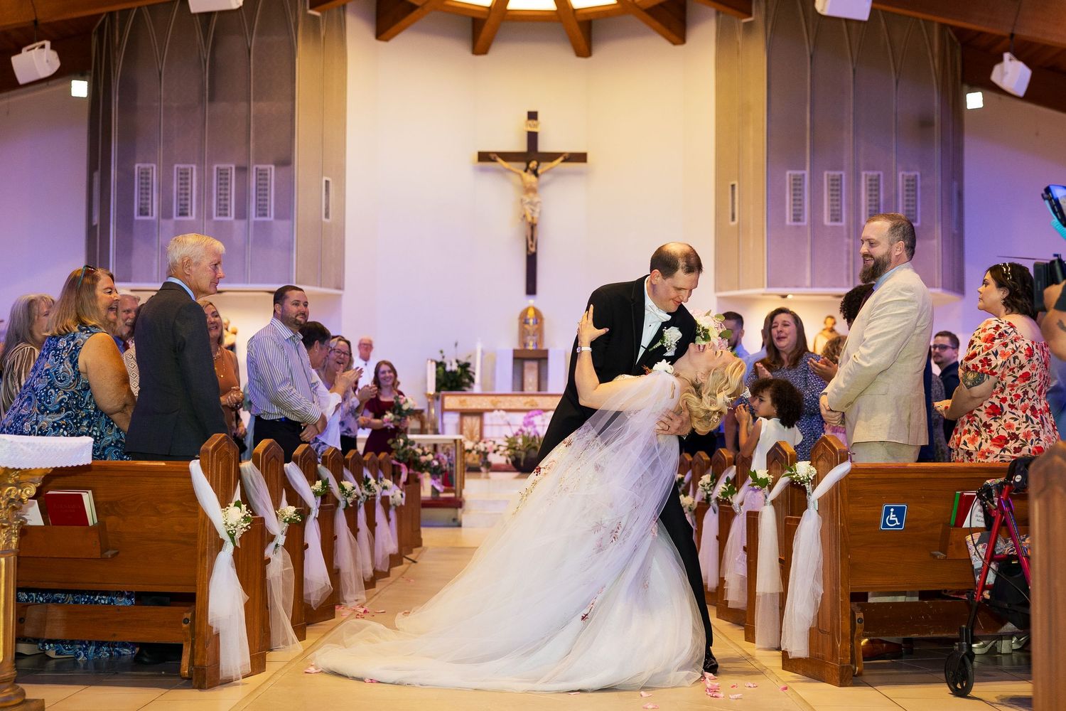 Groom dips bride for romantic kiss in church aisle surrounded by cheering wedding guests.