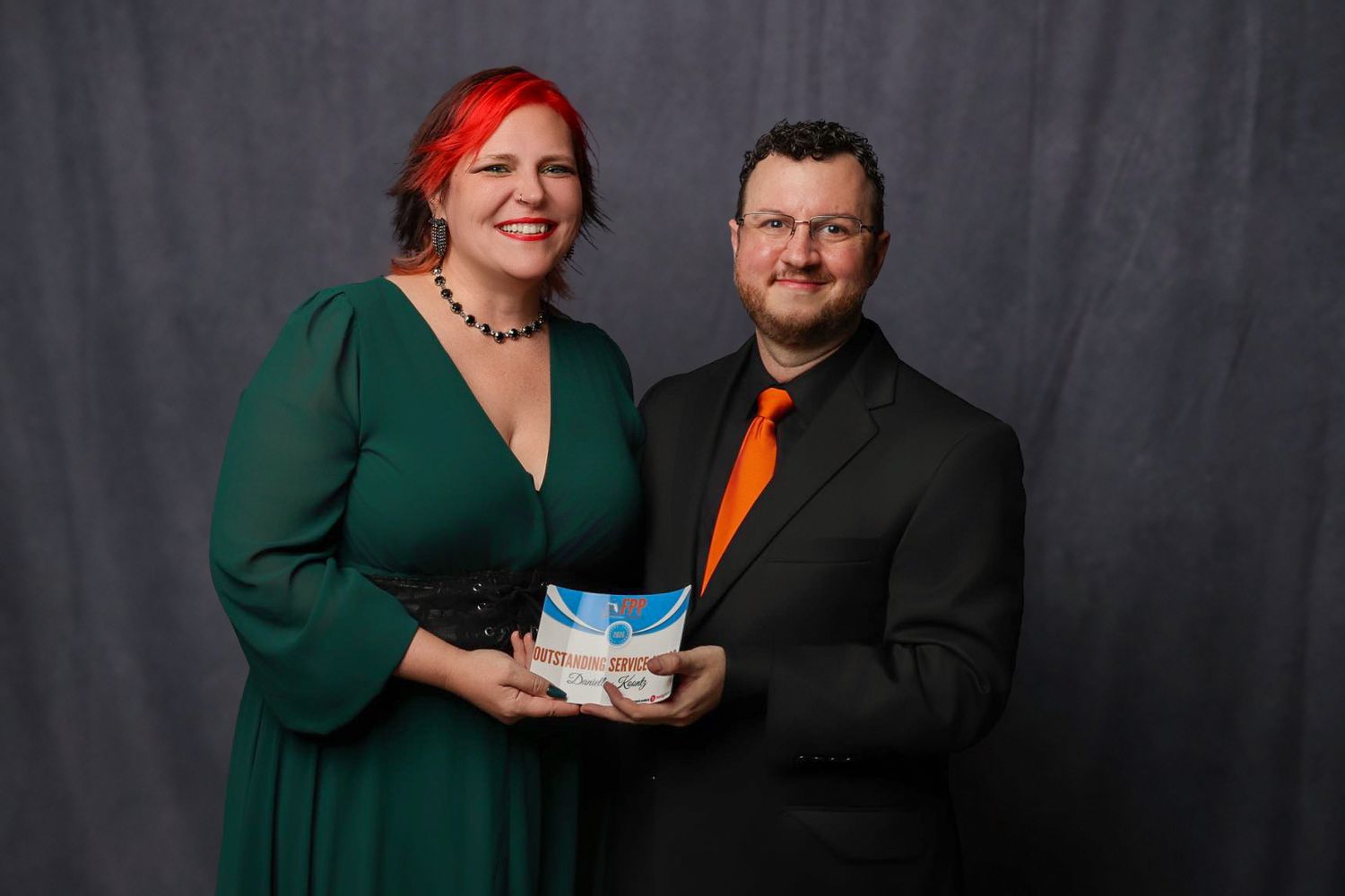Woman with red hair and man in orange tie holding a photography award at a professional ceremony.