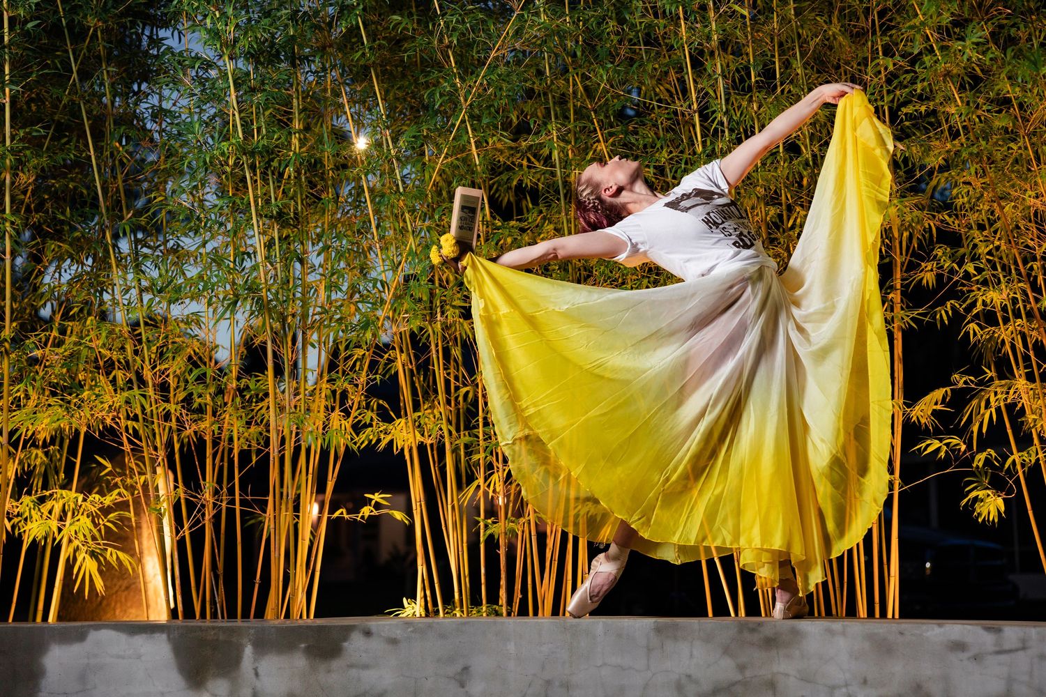 A dancer in a flowing yellow skirt performs an expressive pose among illuminated bamboo stalks at night.