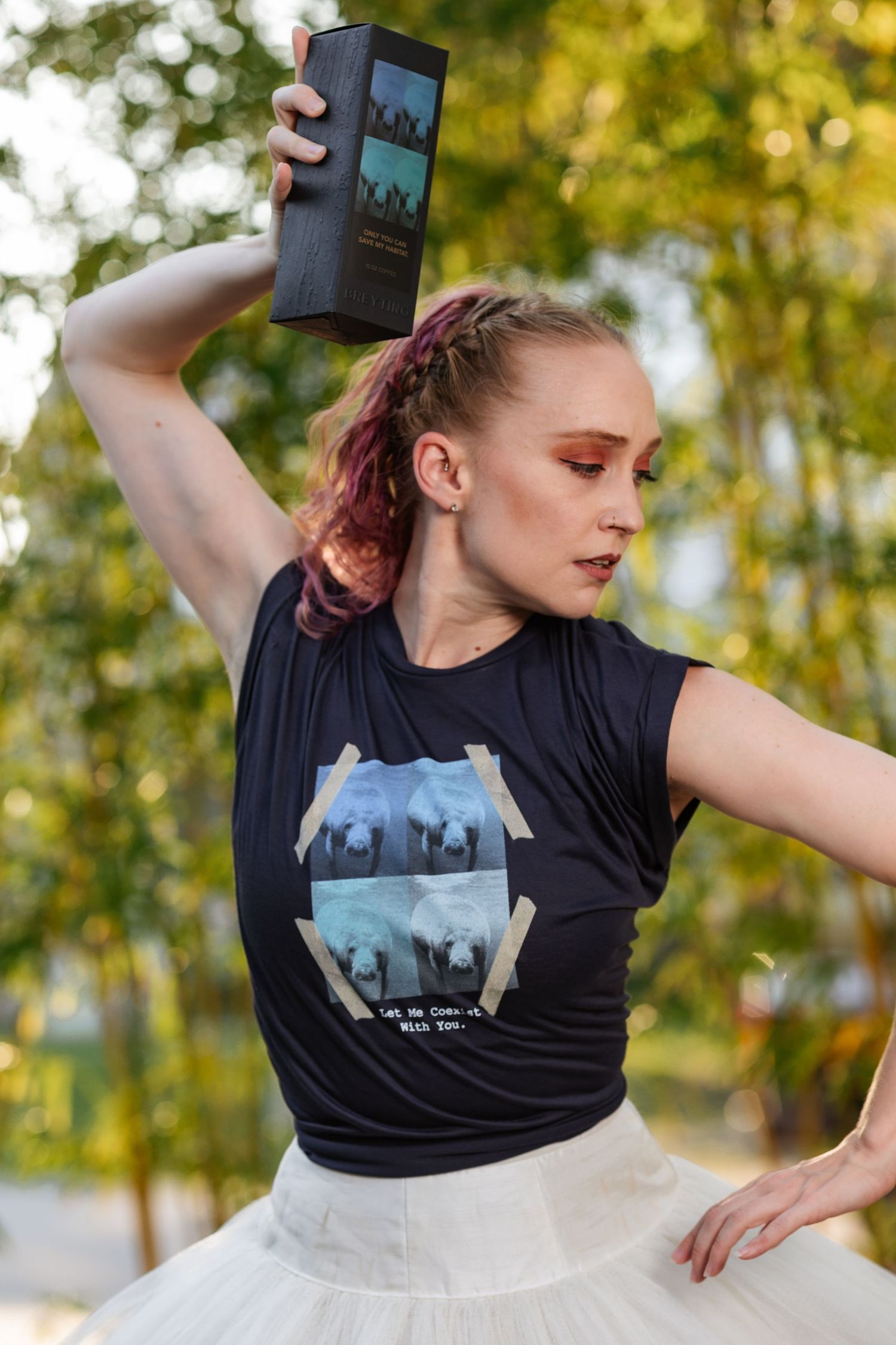 Dancer in black graphic tee and white tutu holding a dark box overhead in an outdoor park setting.