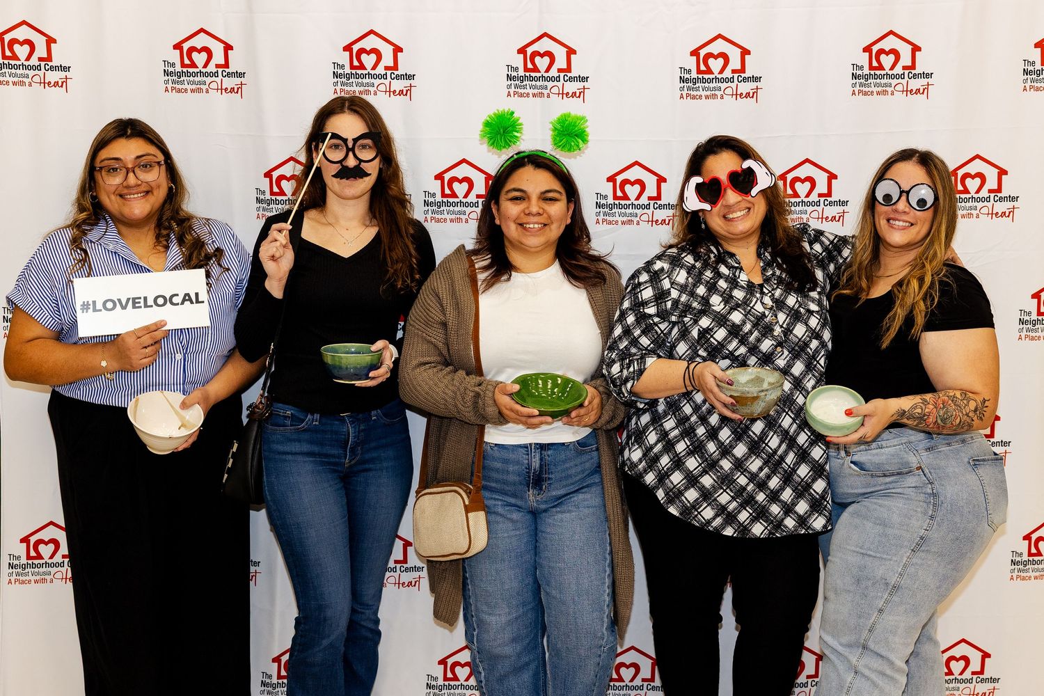 Five women smile and pose with fun props in front of a Neighborhood Housing Fund event backdrop.