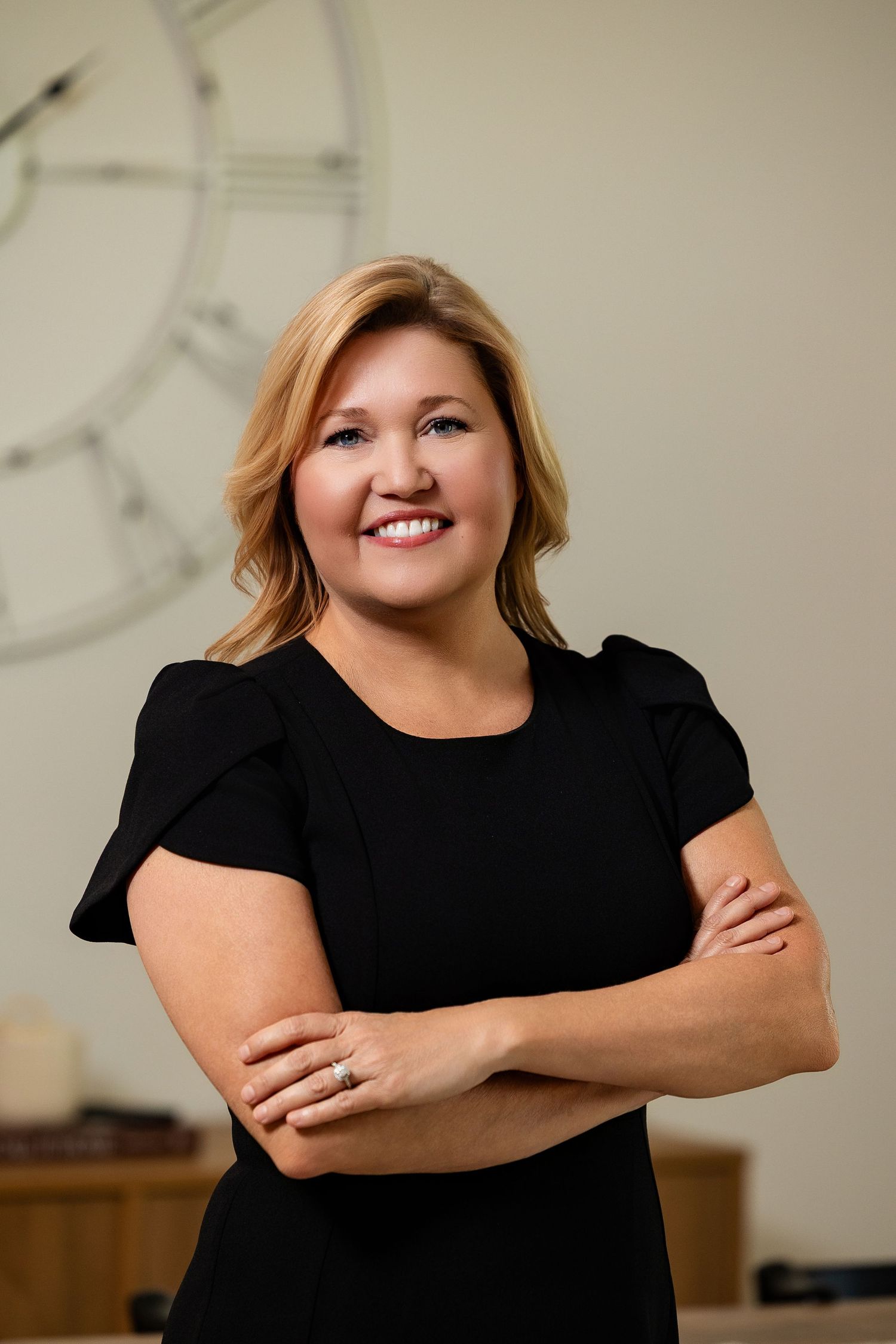 Confident woman in black top with arms crossed smiling in front of decorative clock on wall.
