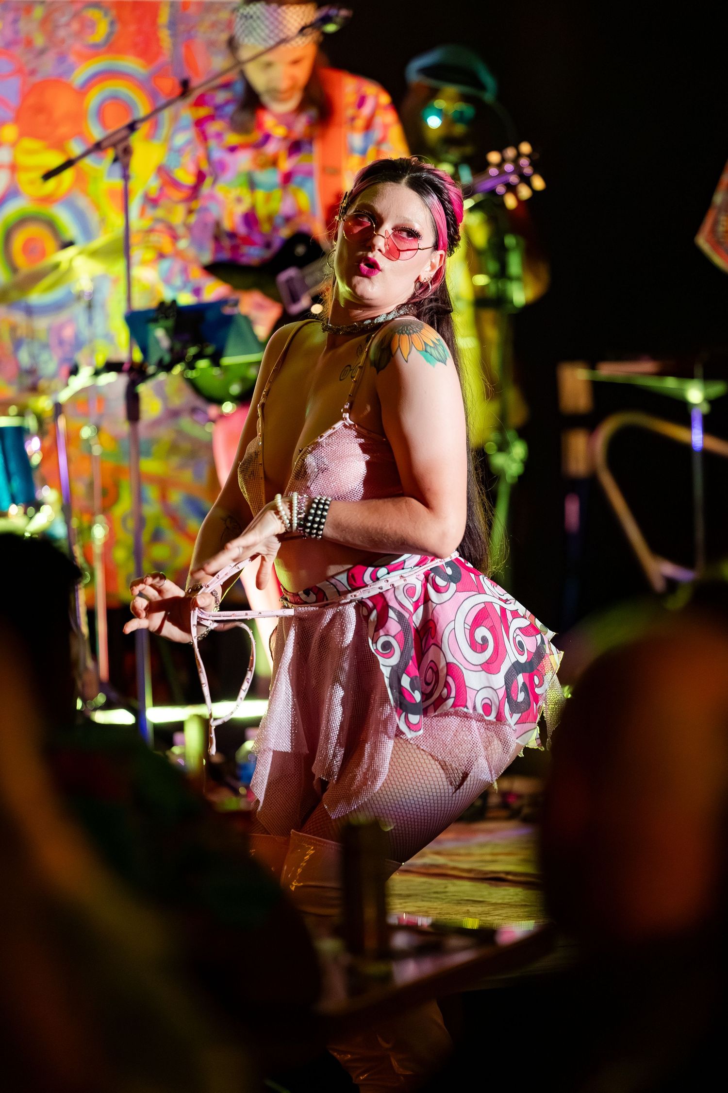 Performer in colorful psychedelic tutu and pink sunglasses on stage at a vibrant retro-themed live show.