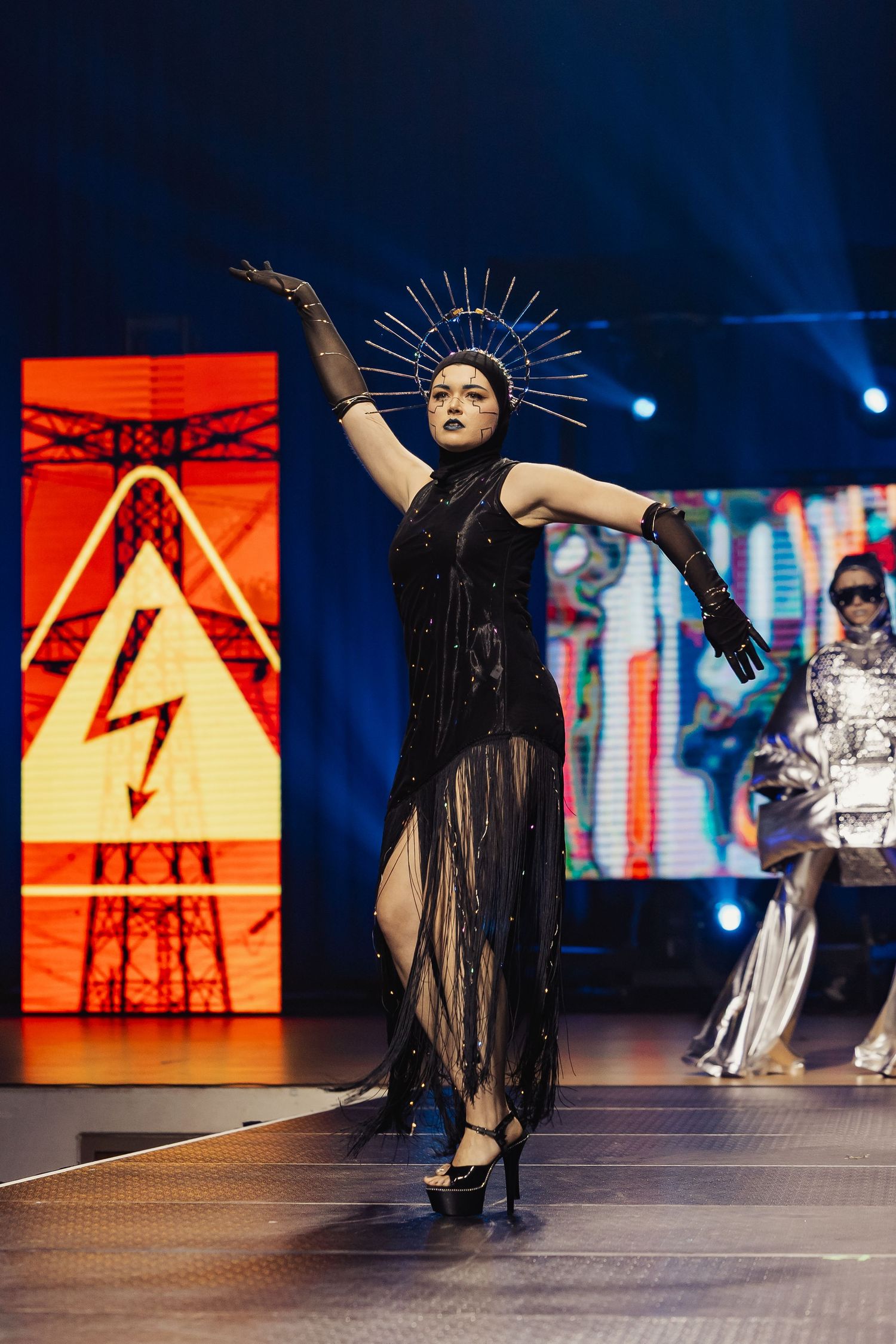 Model in black sequined sheer gown with spiked halo headpiece strikes dramatic pose on runway.
