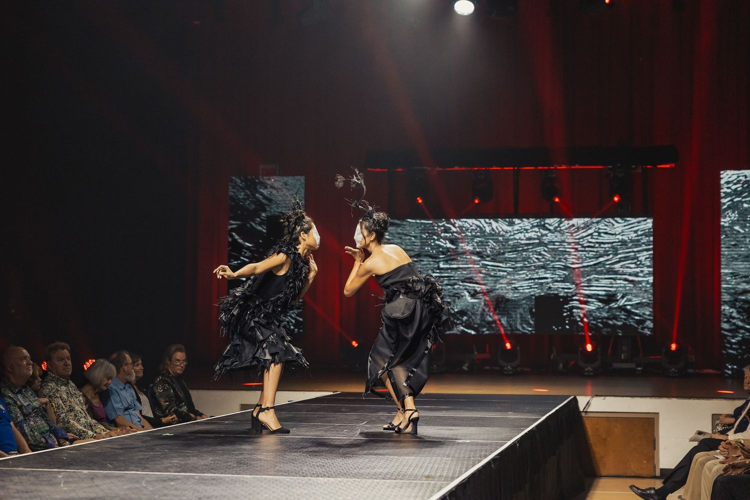Two dancers in dark dramatic costumes perform contemporary dance on stage before a seated audience.