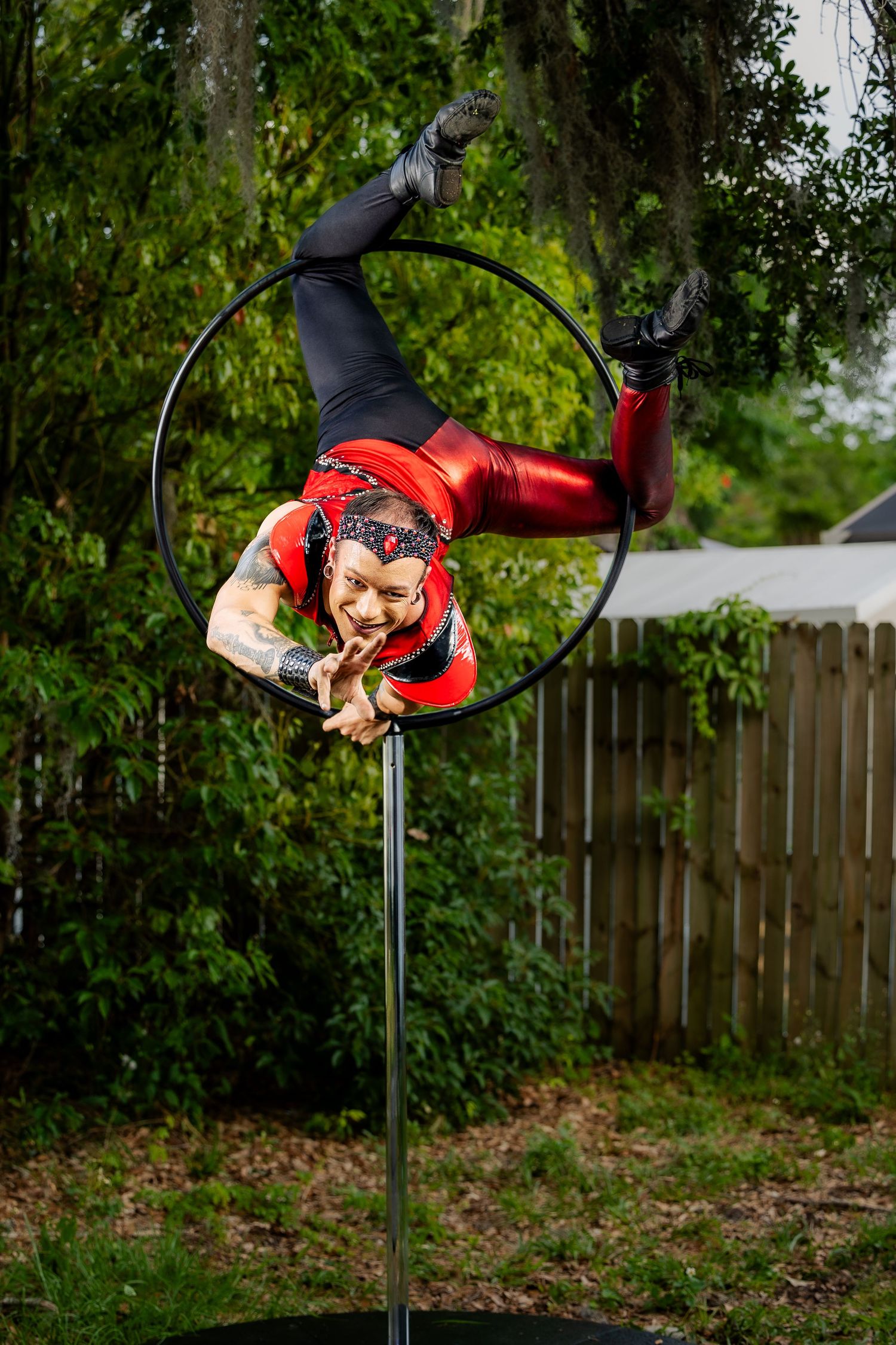 Female aerialist in red and black costume performing a flexible pose inside a lyra hoop mounted on a pole outdoors.