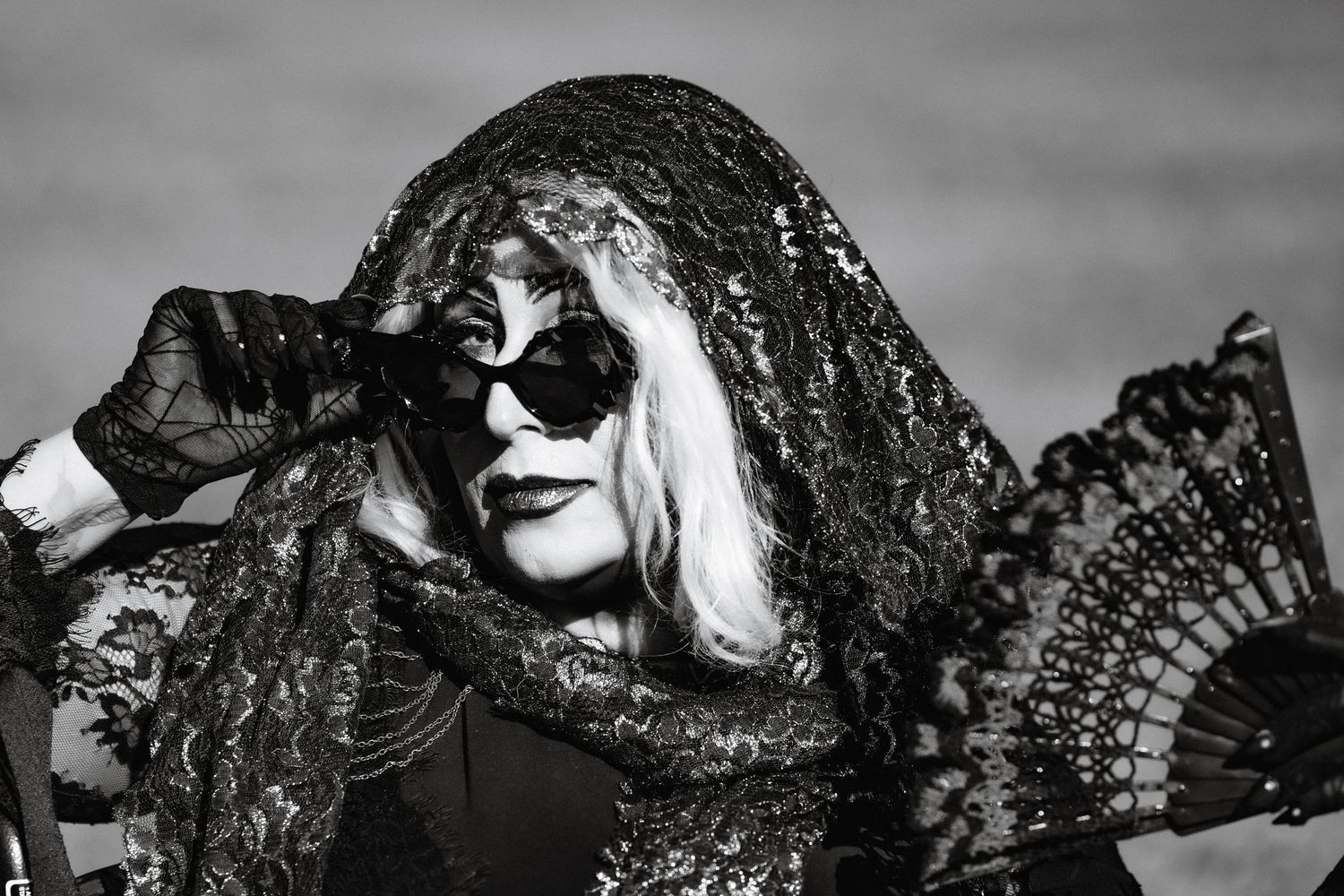 Black and white portrait of woman in ornate black lace veil and mask holding decorative fan.