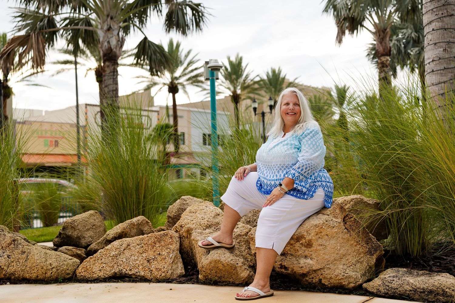 Woman in blue plaid shirt and white pants sitting on rocks surrounded by palm trees and tropical landscaping.