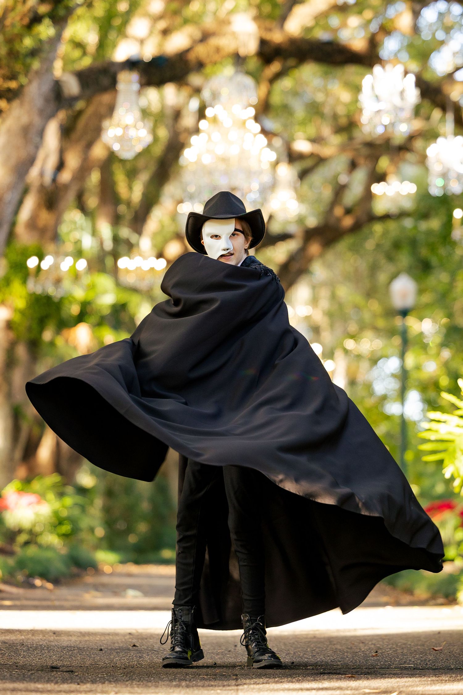 Person in black cape, cowboy hat, and white mask poses dramatically outdoors among trees with chandeliers.
