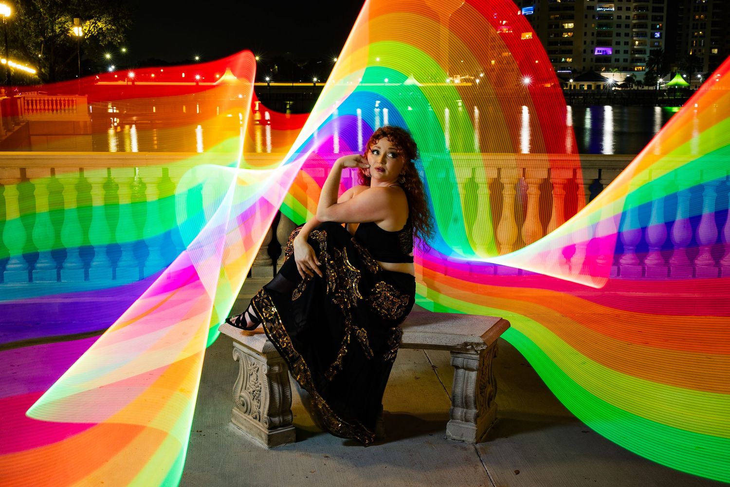 Woman in black dress sits on stone bench surrounded by vibrant rainbow light painting effects at night.