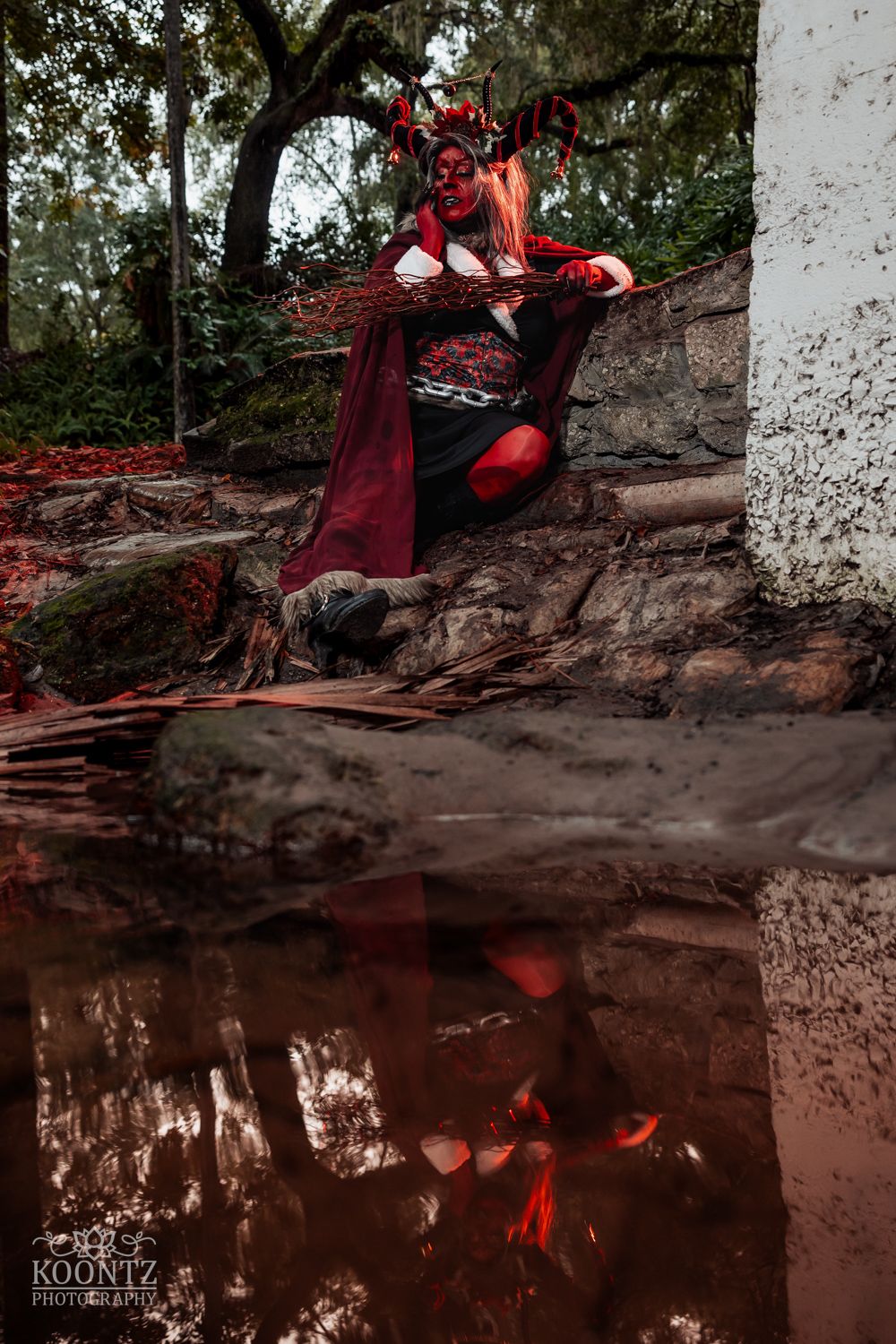 A cosplayer with red face paint, antler headpiece, and red cape sits by a reflective pool in a forest setting.