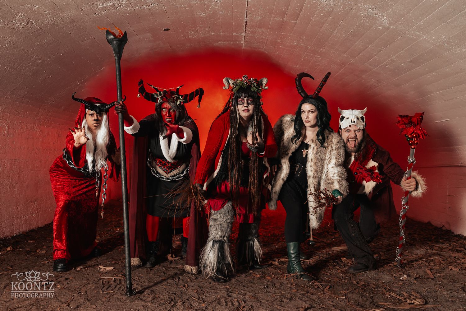Four cosplayers in red and black devil costumes crouch and pose in a tunnel with red glow backdrop.