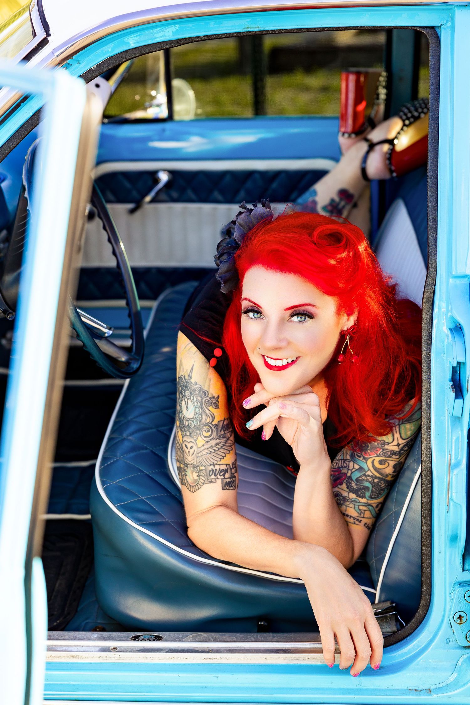 Red-haired tattooed pinup woman smiling inside vintage blue classic car interior.