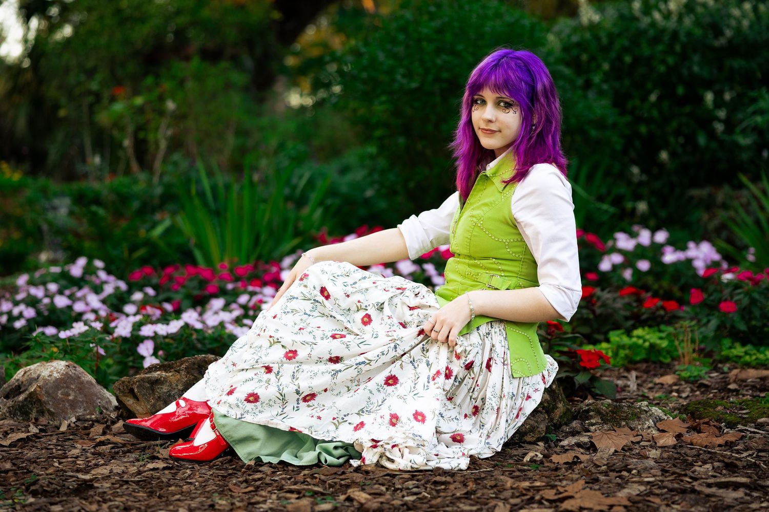 Woman in purple wig, floral skirt, and green vest sits among colorful garden flowers in cosplay outfit.