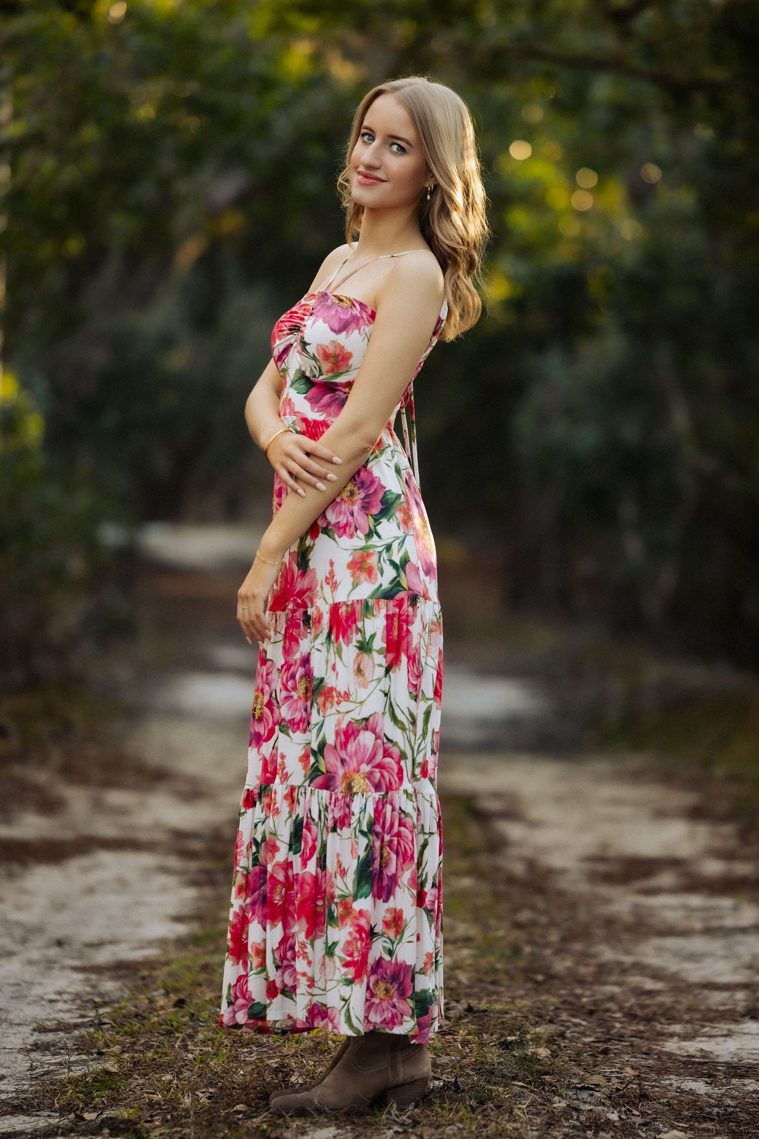 Young woman in floral maxi dress standing on a dirt path surrounded by lush green trees.
